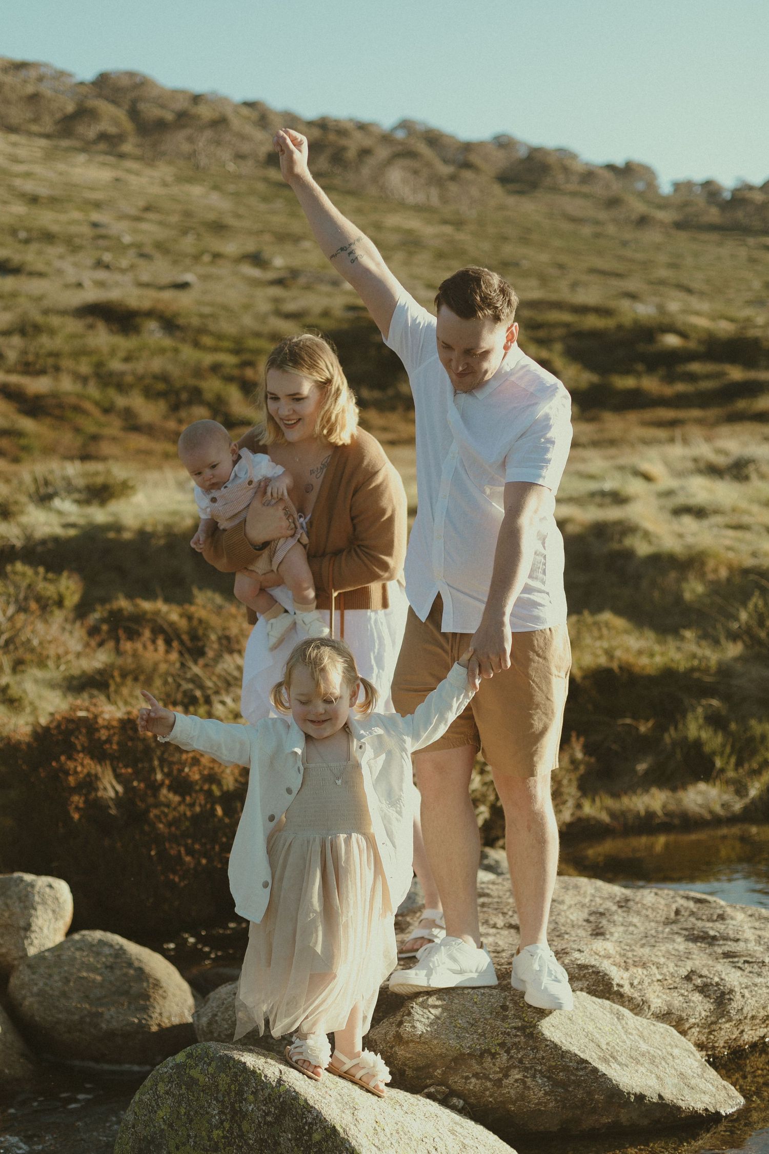 Family in coordinated neutral outfits poses together on rocky terrain during golden hour sunset.