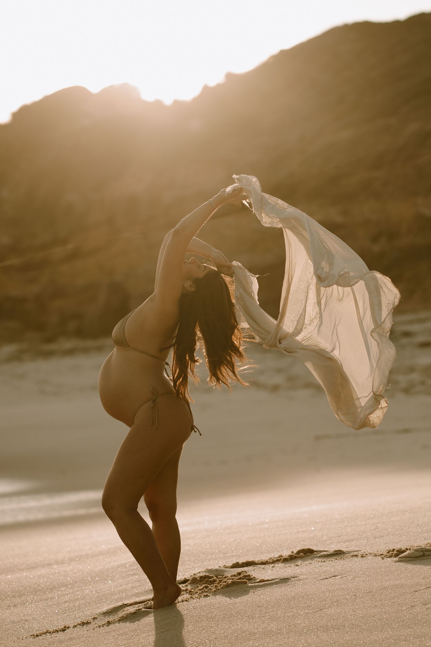 Silhouetted dancer arches backwards on beach at sunset while fabric billows dramatically in golden light.