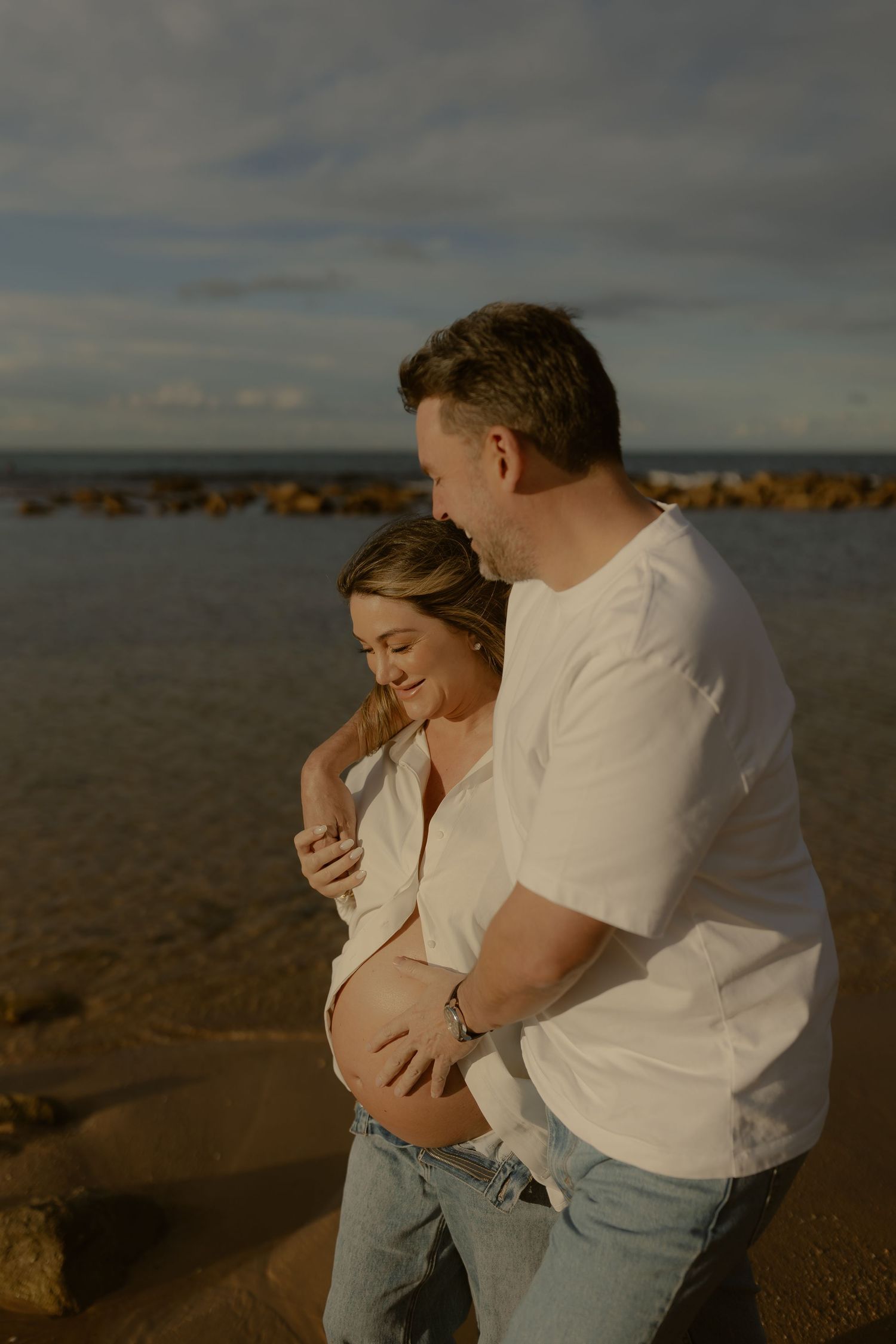 Couple embracing on beach at golden hour, man holding pregnant womans baby bump near ocean rocks.