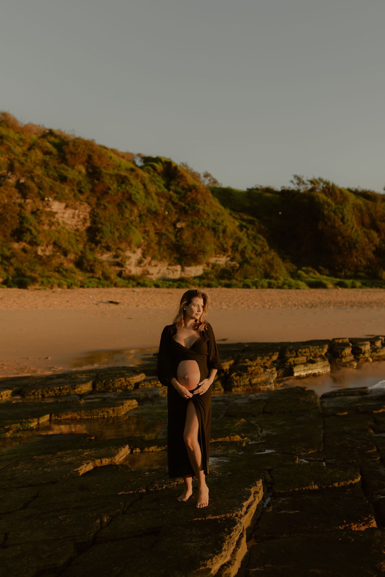 Pregnant woman in black dress cradling baby bump on rocky beach at golden hour sunset.