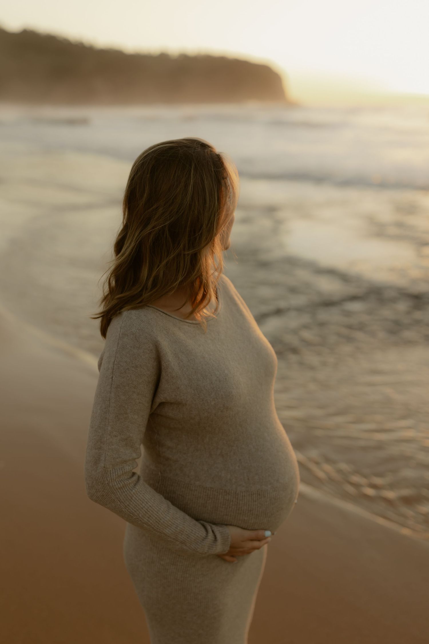 Pregnant woman in beige knit dress holding bump while gazing at golden sunset beach waves.