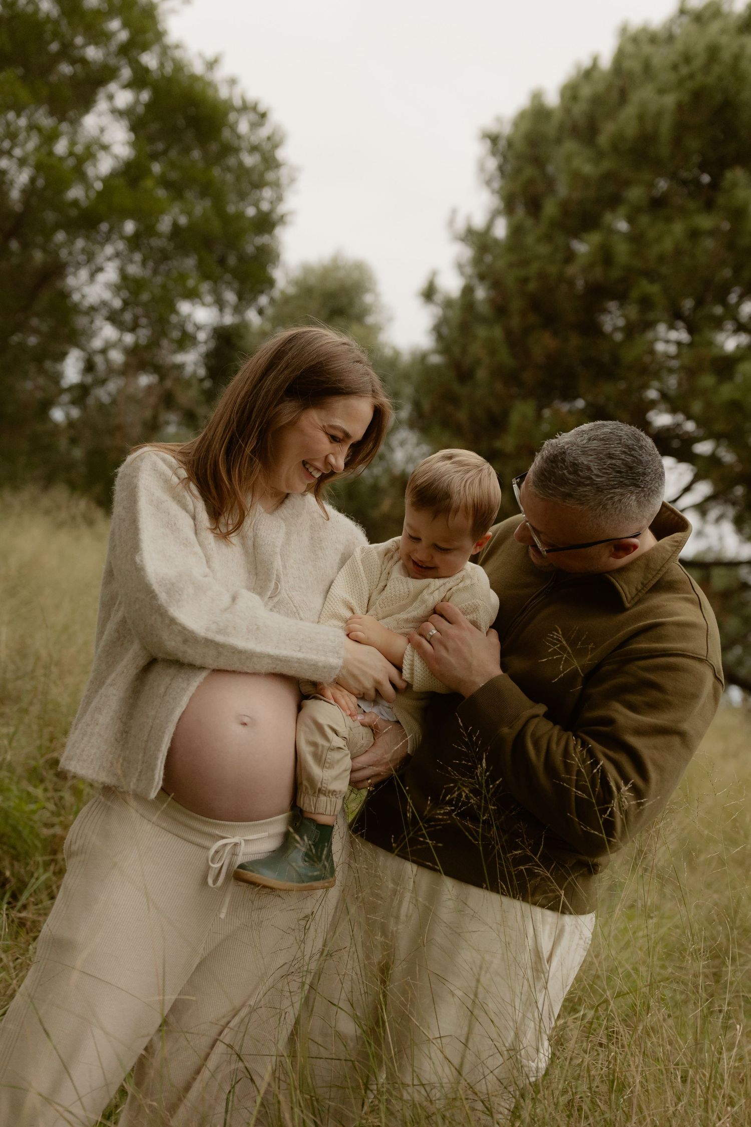 Smiling pregnant couple in neutral tones hold their toddler son together in a lush green field.