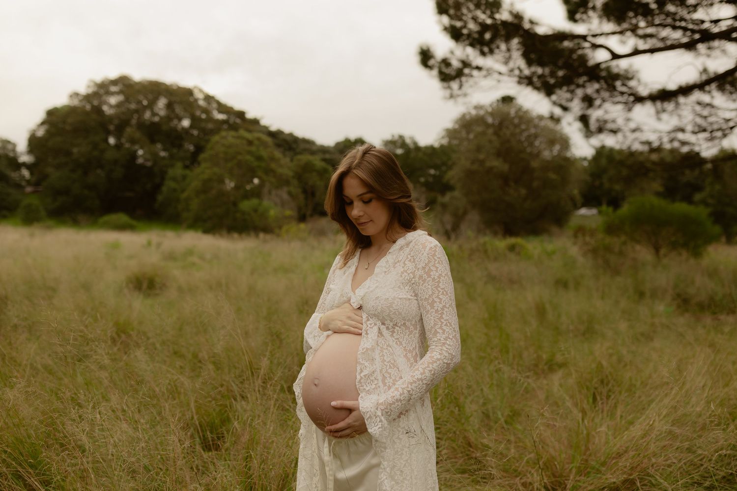 Pregnant woman in white lace dress cradling baby bump in a serene grassy field.