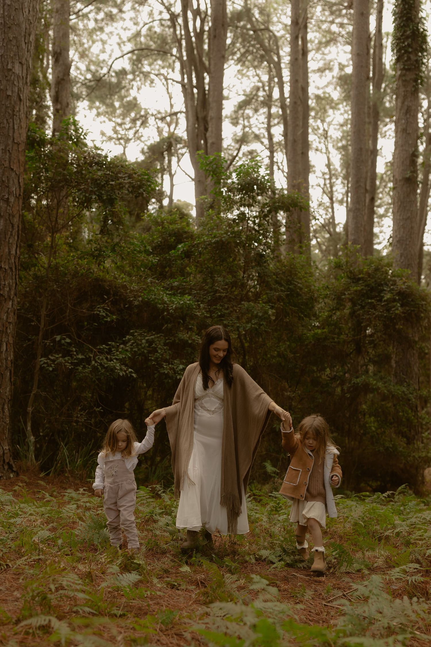 Mother in white dress walks through forest holding hands with two young children.