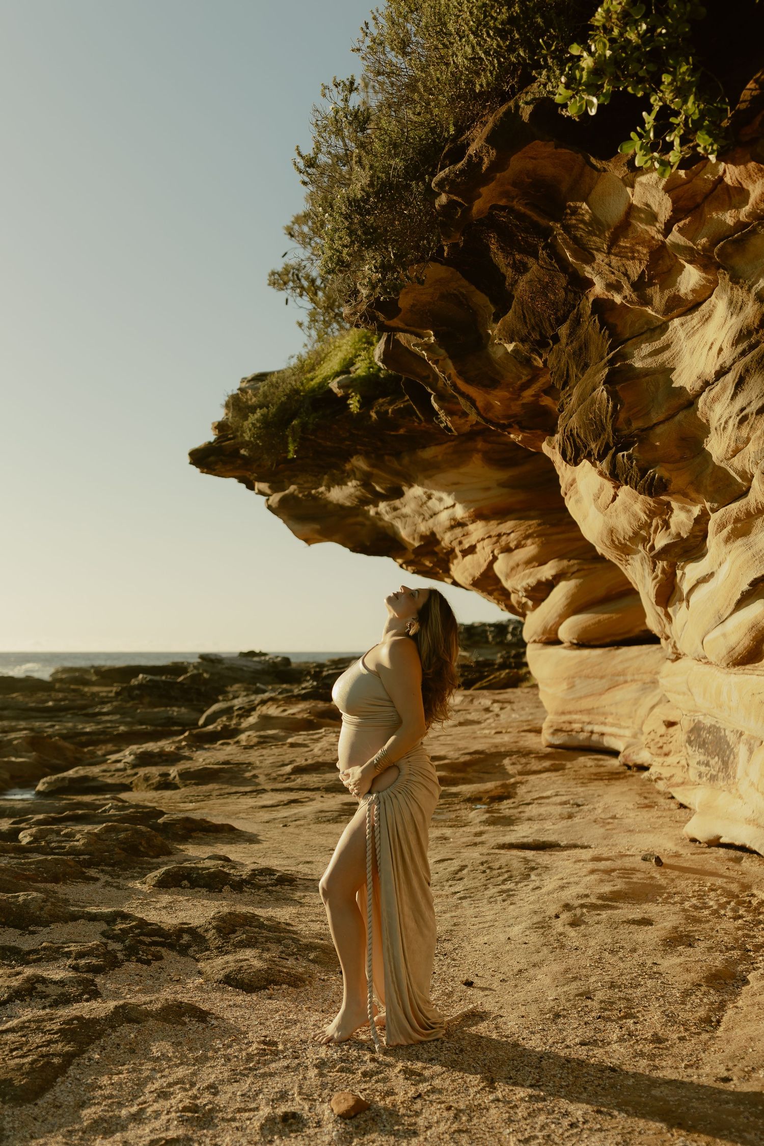 Pregnant woman in flowing dress poses by golden sandstone cliffs at a rocky beach during golden hour.