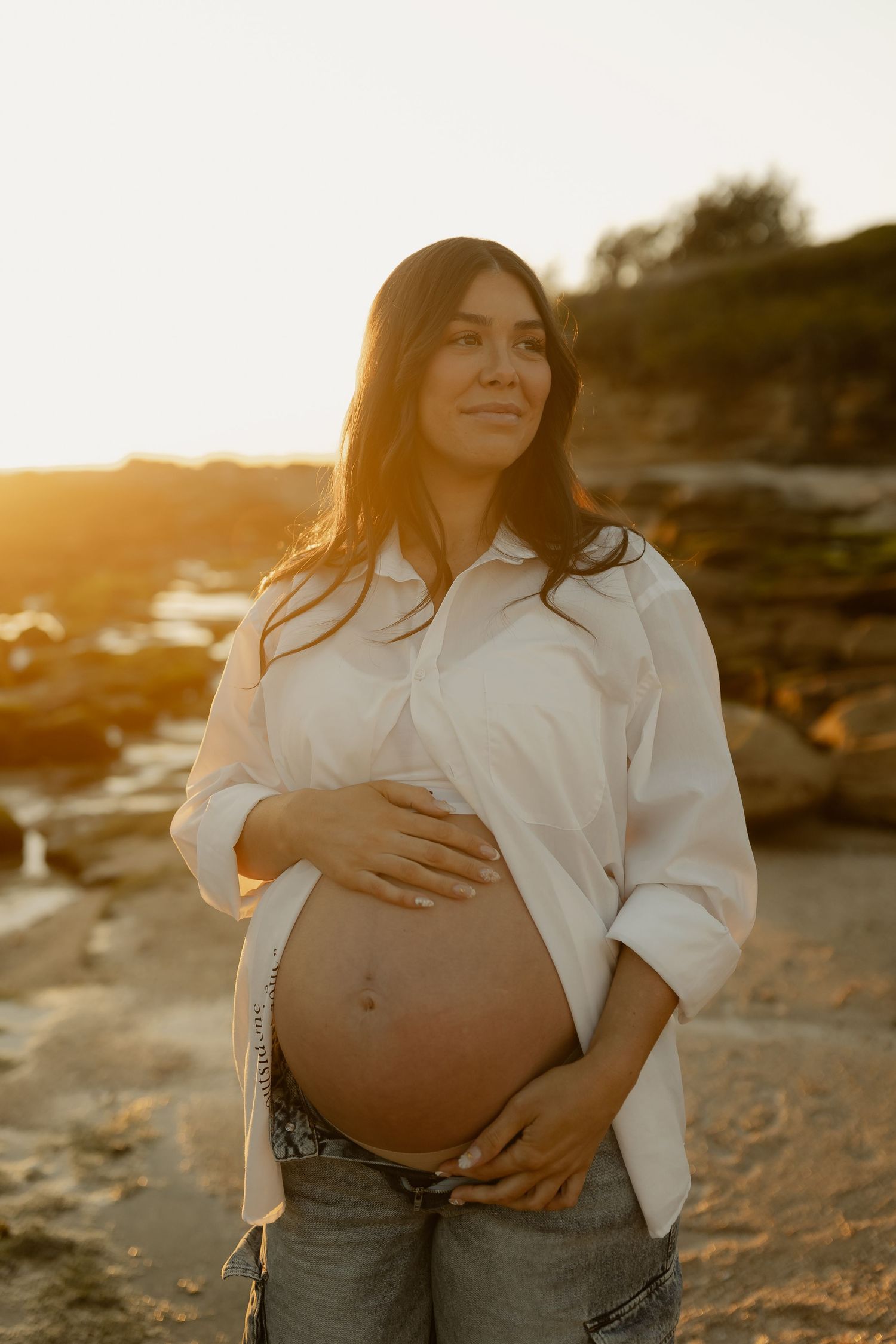 Pregnant woman in white shirt holding baby bump on rocky beach during warm golden sunset.
