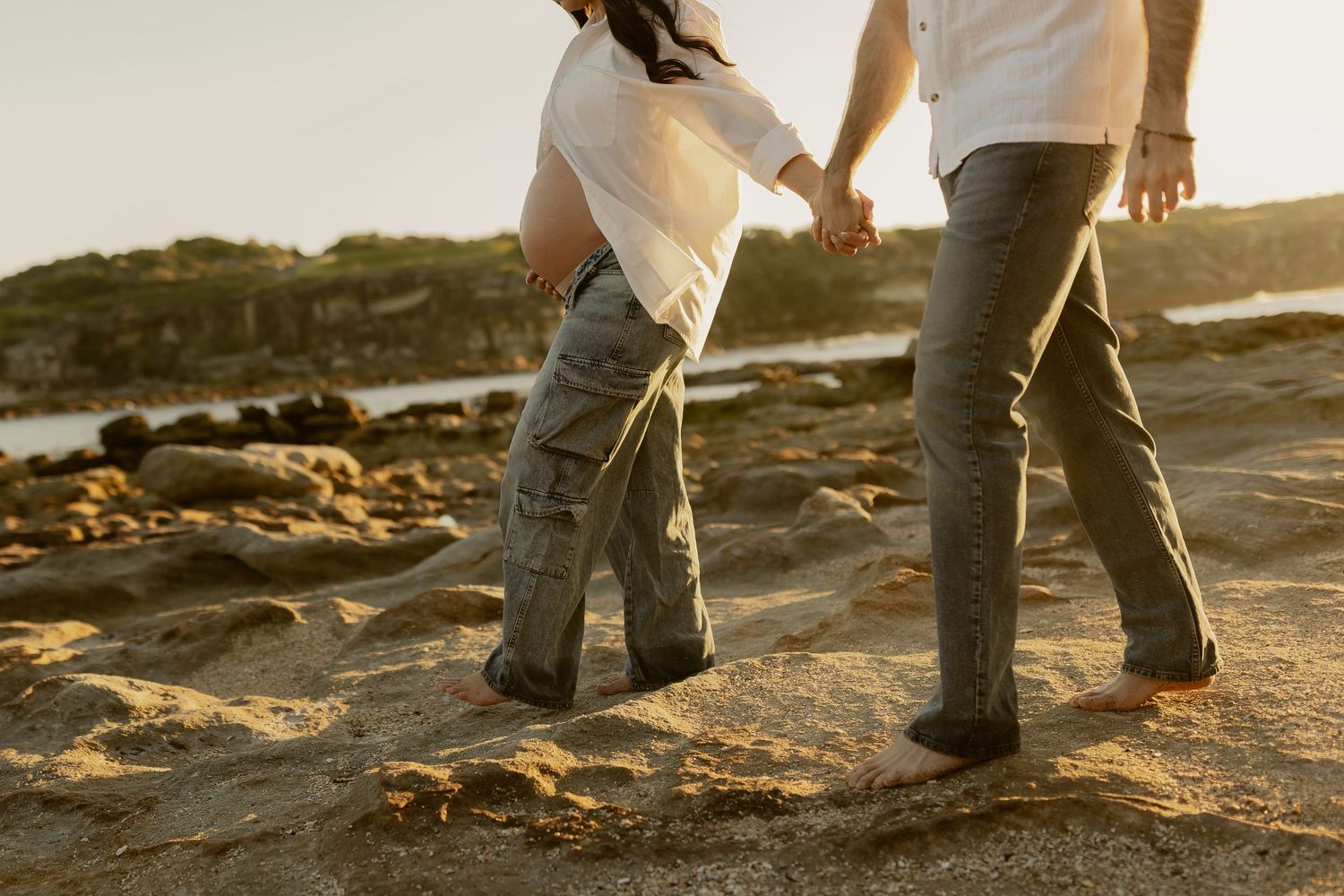 Couple walking barefoot on rocky beach at golden hour, wearing white tops and gray pants.