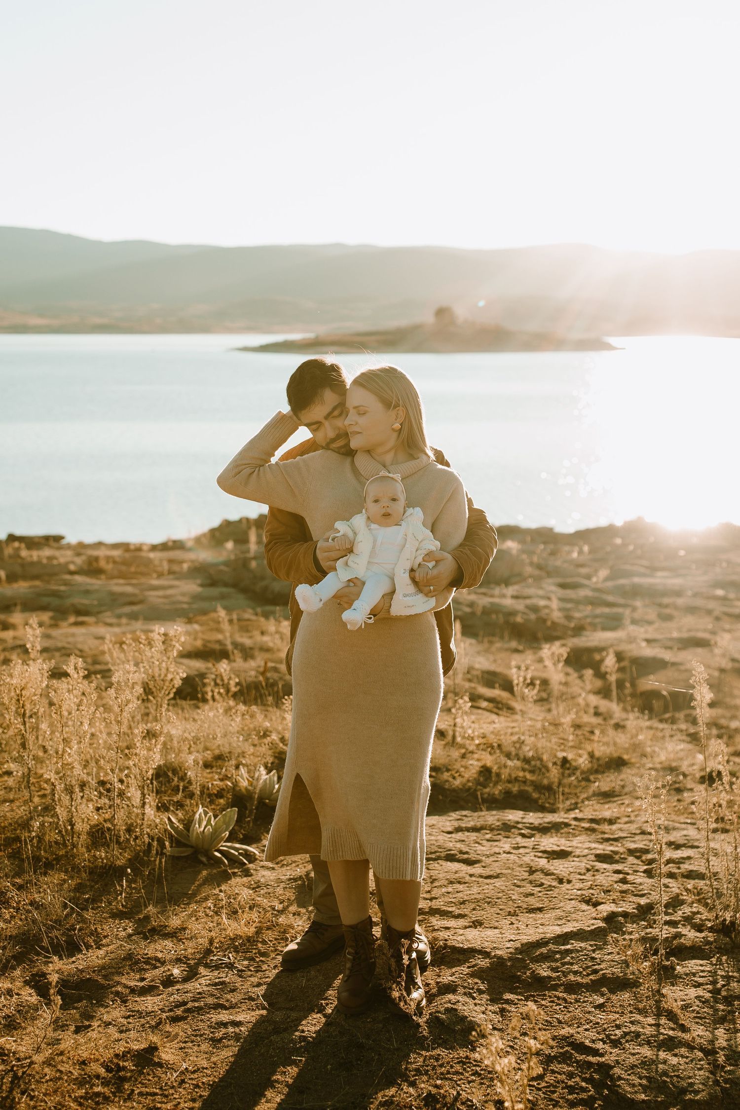 A tender family moment captured in golden sunlight by a lakeside landscape.