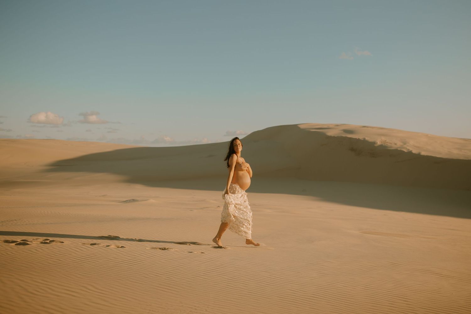 A lone figure walks across golden sand dunes in the desert during sunset, wearing flowing white clothing.