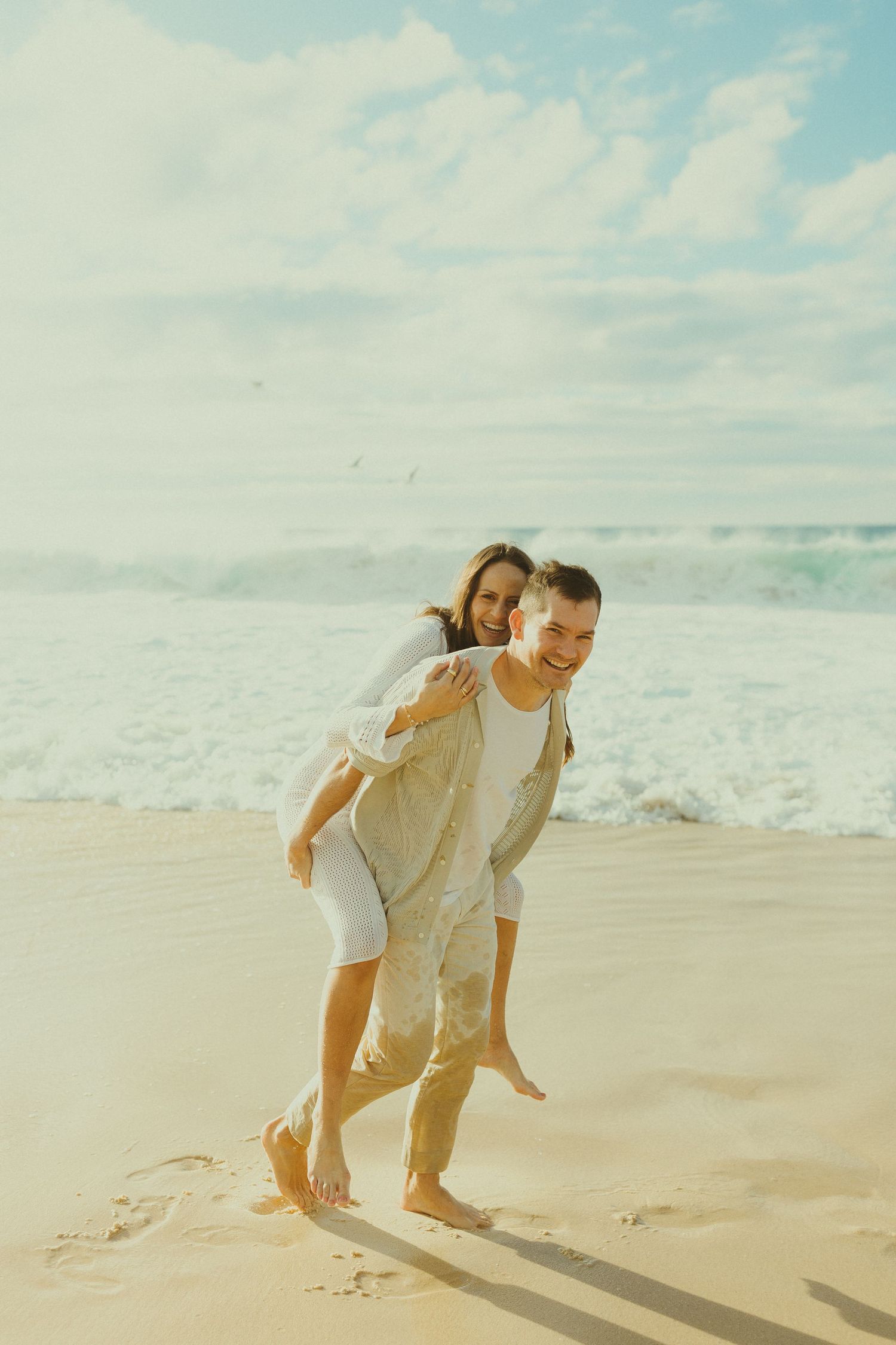 A couple enjoying a playful moment together on a sunny beach with waves and blue sky in the background.