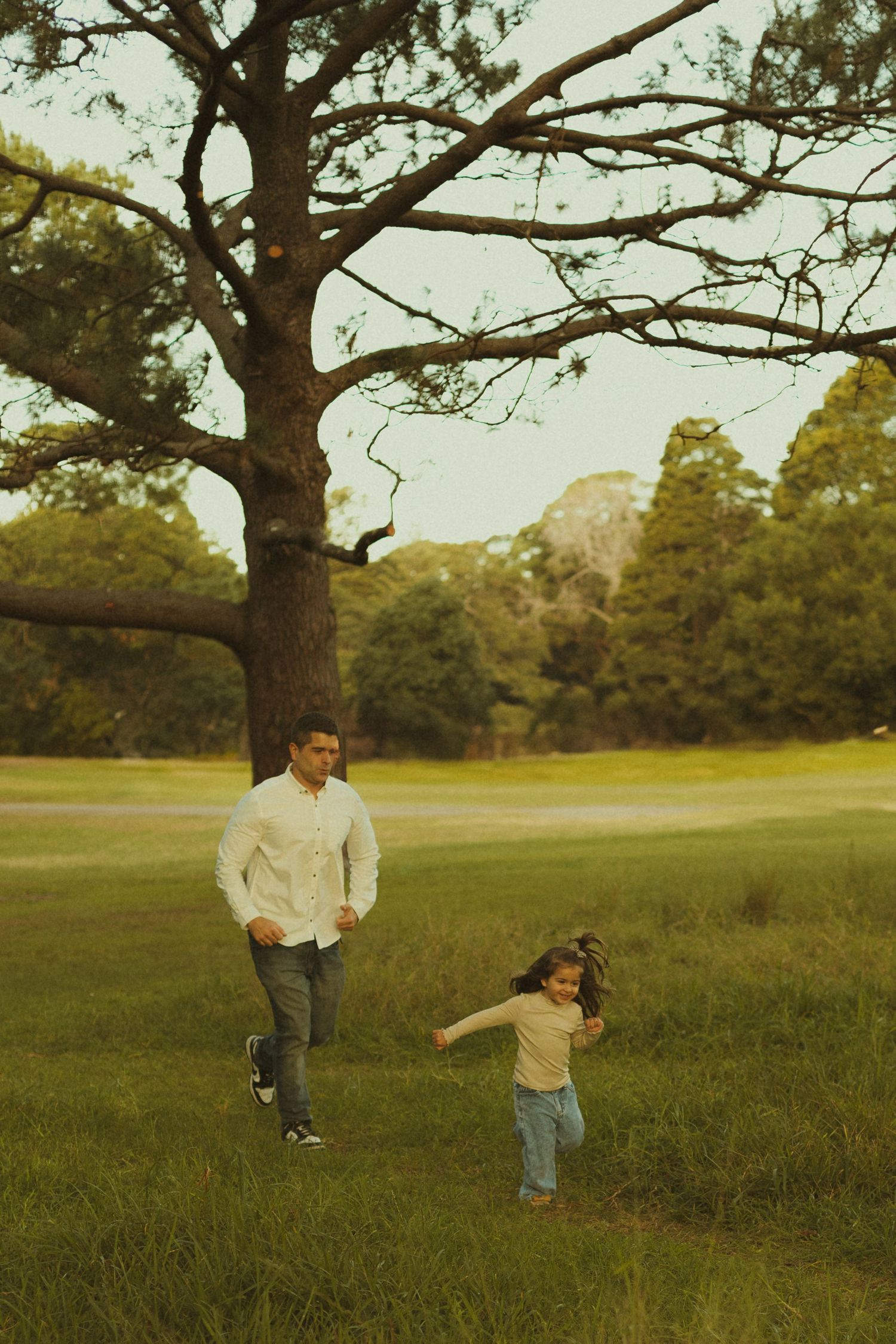 Two people run playfully through a grassy field under a large tree branch on a warm autumn afternoon.