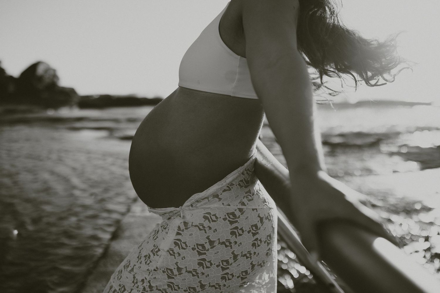 Black and white photo of pregnant woman in floral dress walking on beach during sunset with wind blown hair.