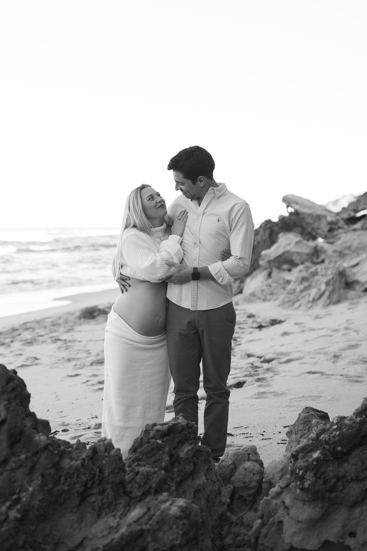 A black and white beach portrait features an expecting couple standing among coastal rocks at sunset.