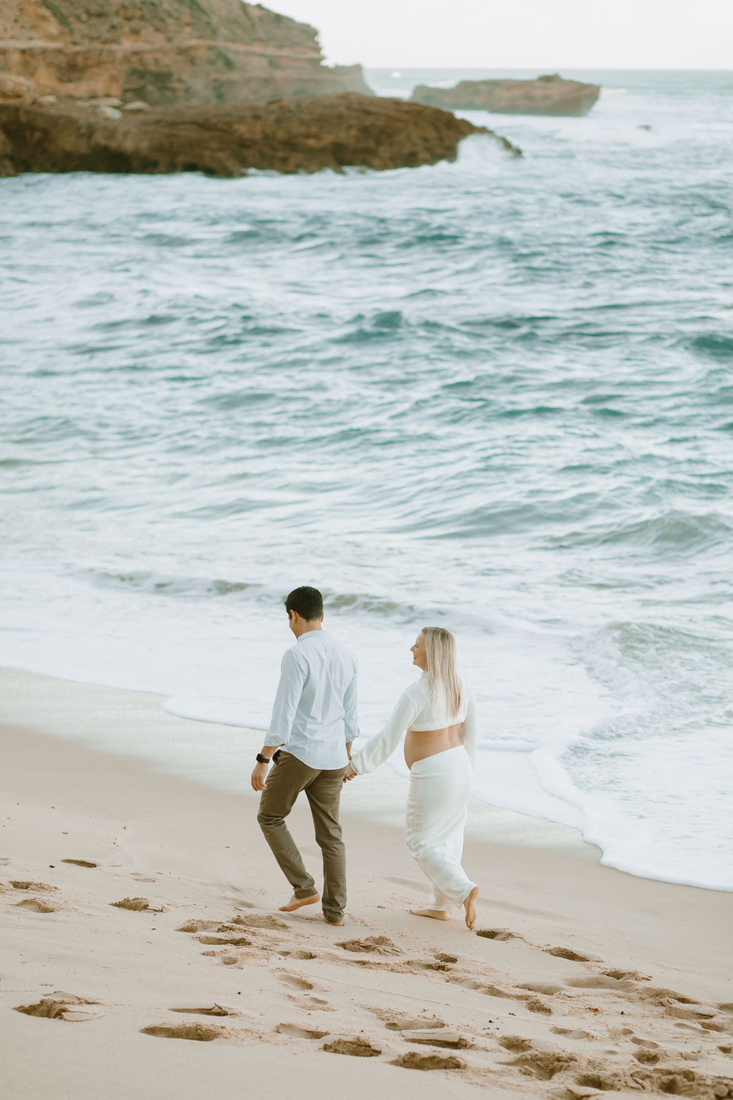 Two people walk hand in hand along a scenic beach with rocky cliffs and crashing waves in the background.