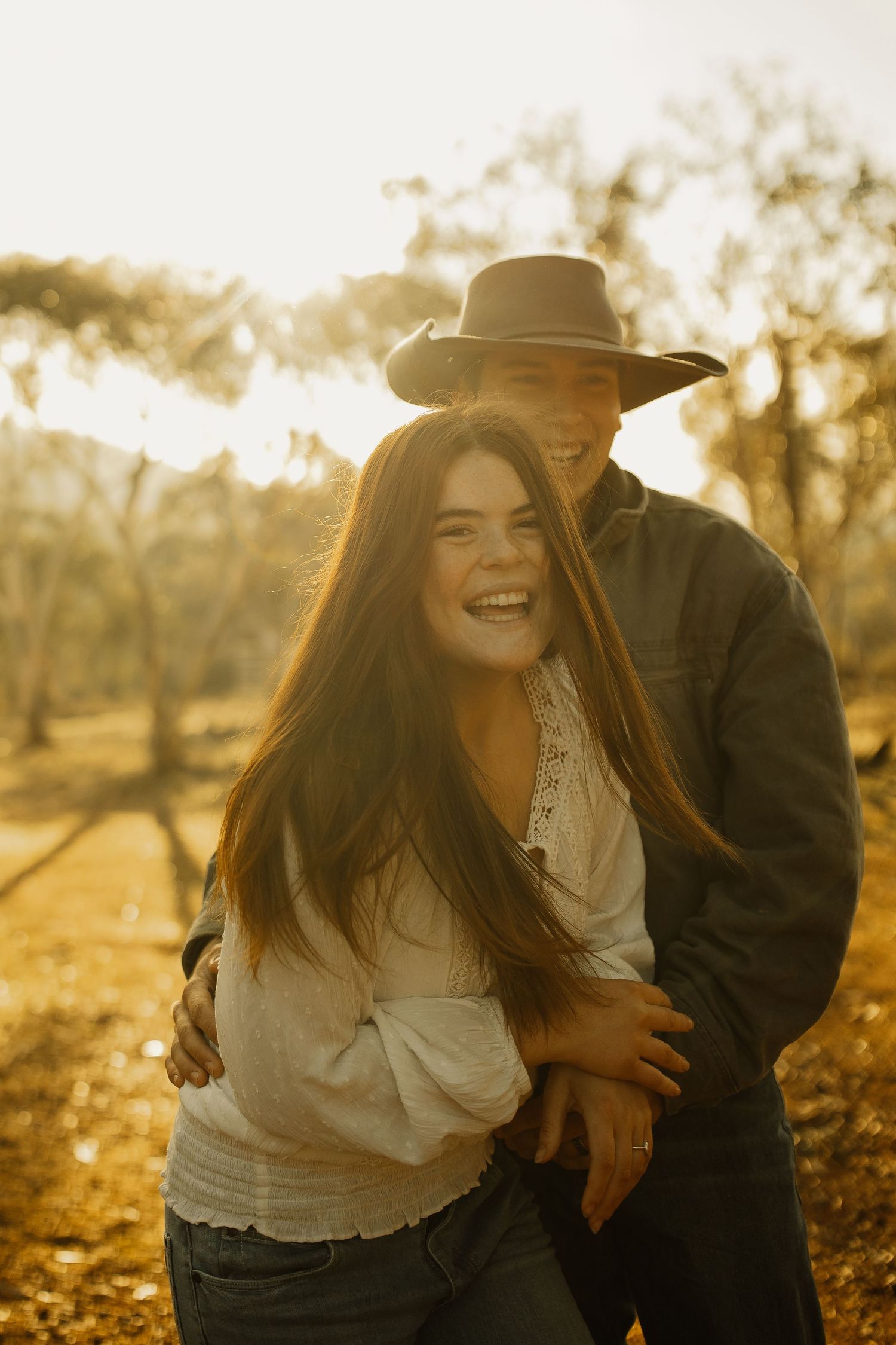 A romantic couple shares laughter in warm sunset light while embracing outdoors in casual autumn attire.