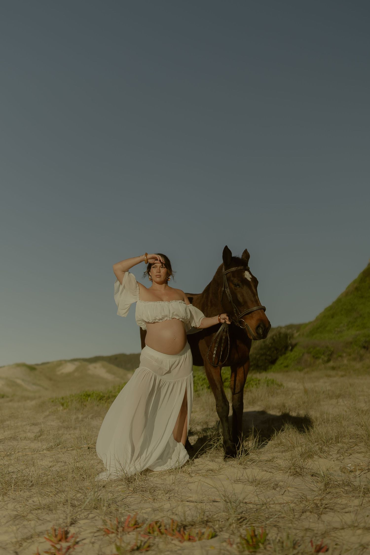 Pregnant woman in white off-shoulder dress poses with brown horse on sandy dunes under clear sky.