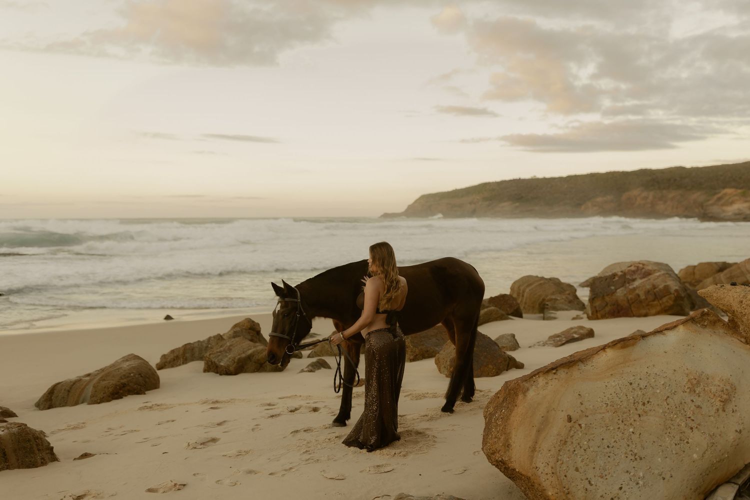 A person stands with a dark horse on a sandy beach at sunset, with rocky shores and ocean waves in the background.