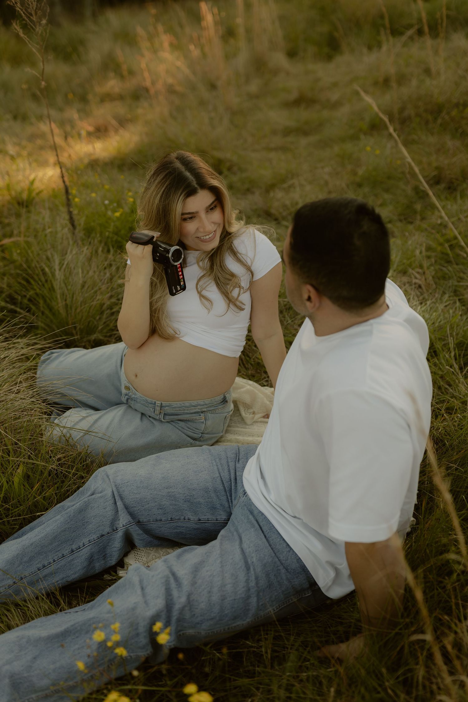 Pregnant couple sits in grassy field at sunset, woman holds vintage camera smiling.