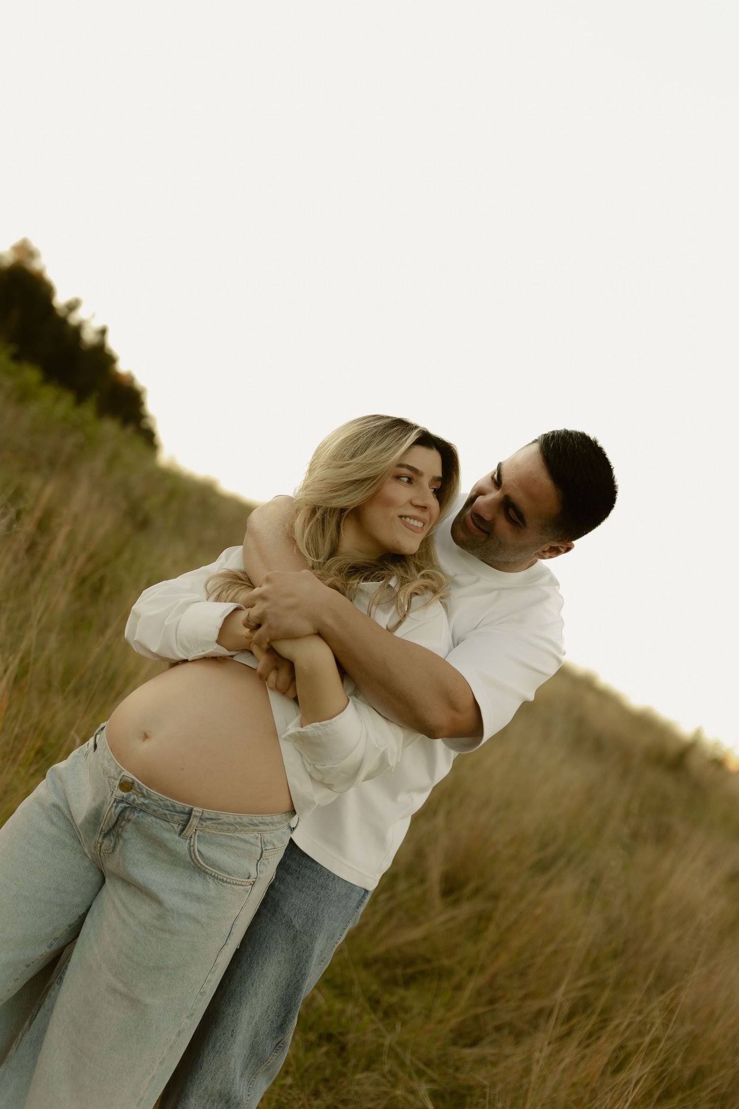 A couple embraces outdoors in a golden field, the woman visibly pregnant, both wearing white tops and jeans.