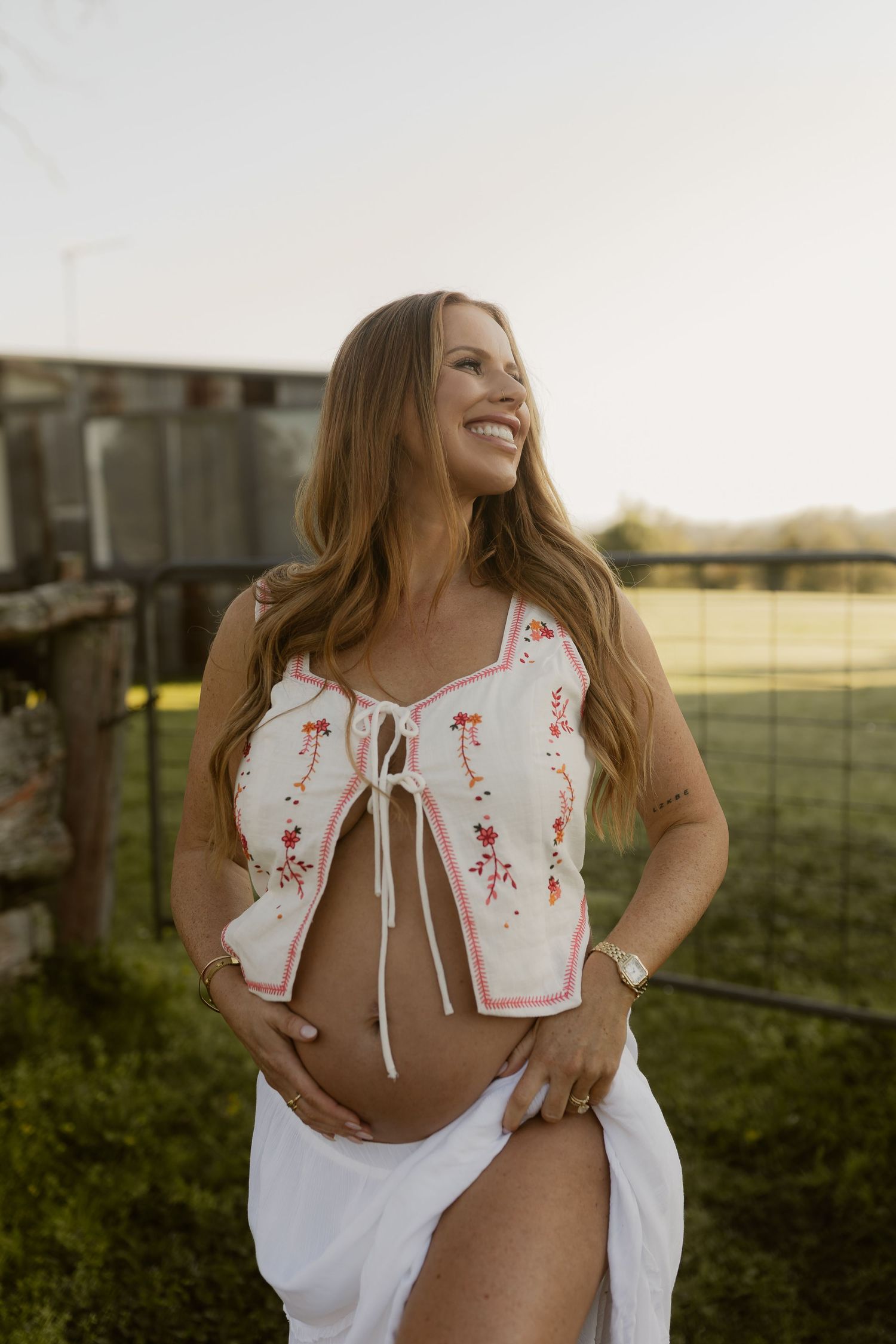 Smiling pregnant woman in white floral crop top and skirt cradling baby bump outdoors near a fence at golden hour.