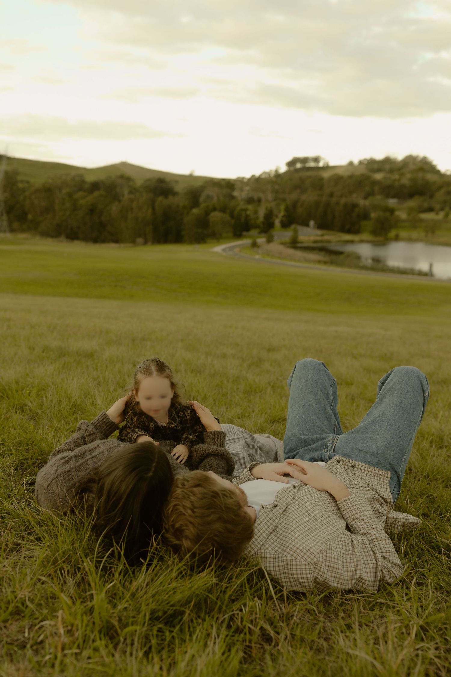 Family relaxing on lush green grass in a scenic countryside with rolling hills and a pond at dusk.