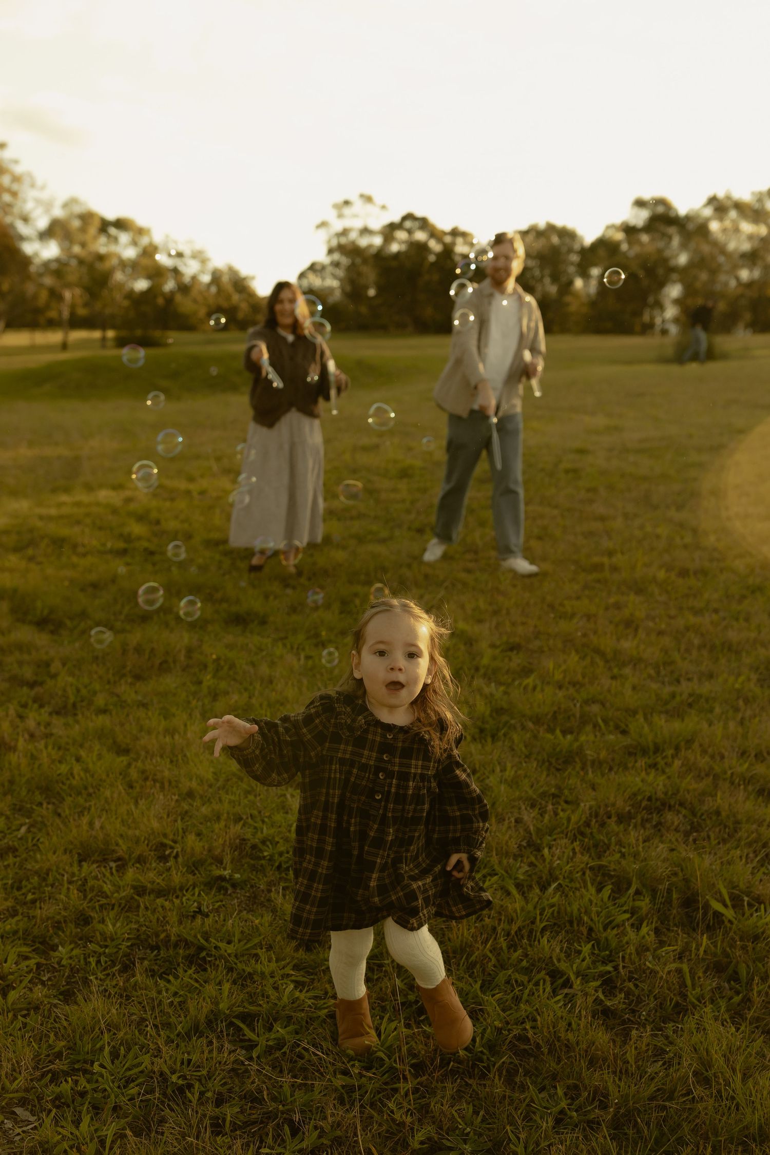 A young girl in a plaid dress chases bubbles in a sunny park while her parents blow more bubbles behind her.