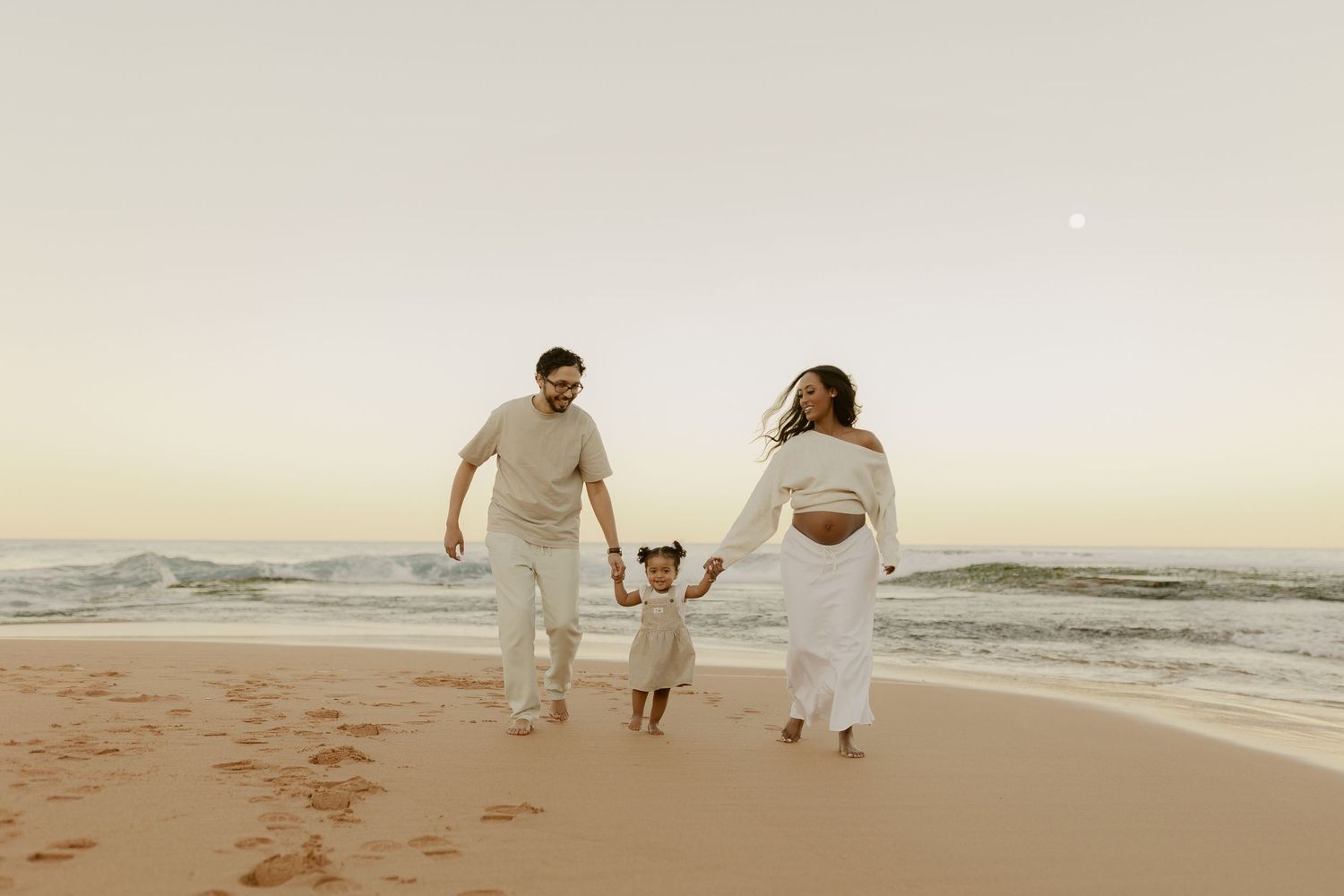 Family in white outfits walking on sandy beach at sunset, parents holding toddler's hands near ocean waves.