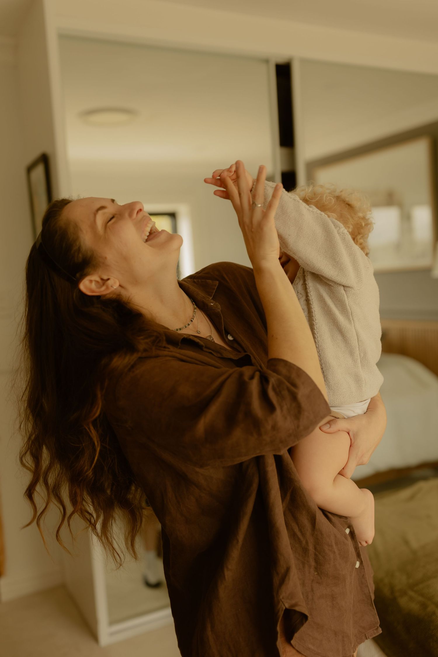 A laughing woman in a brown linen shirt joyfully lifts a baby in a bright, modern bedroom.