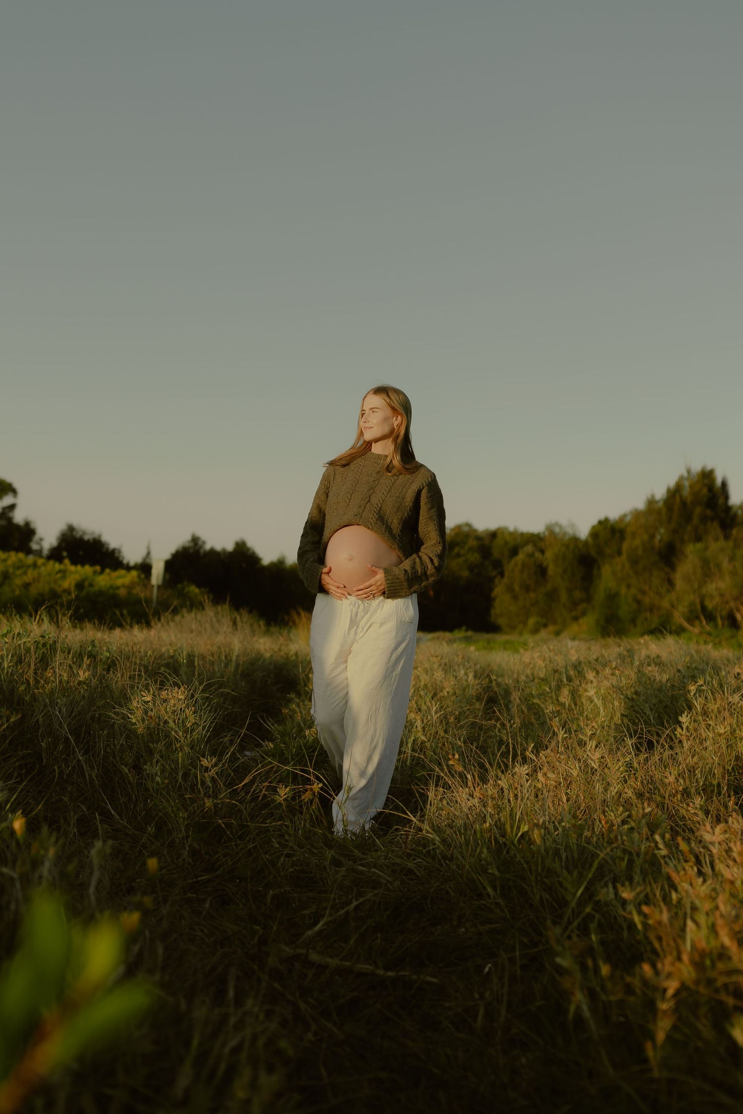 Pregnant woman in green sweater and white pants standing in a sunlit meadow at golden hour.