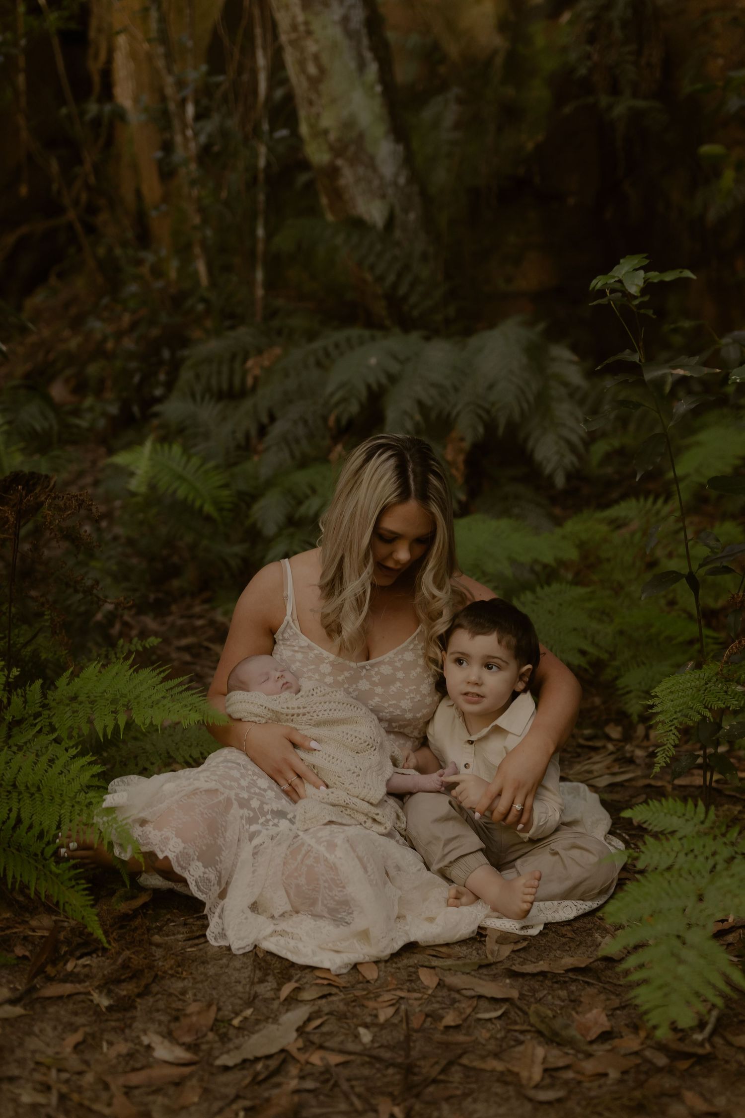 Mother in floral lace dress sits on forest floor with toddler son while gently holding newborn baby.