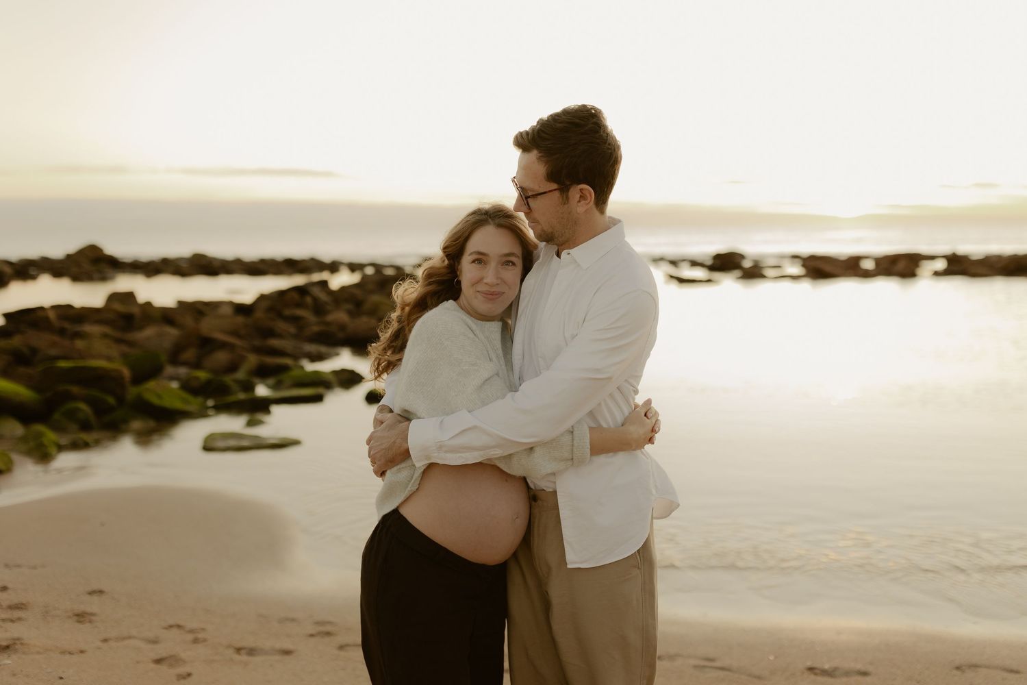 Couple embracing on beach at sunset, pregnant woman cradled by partner near rocky shoreline.