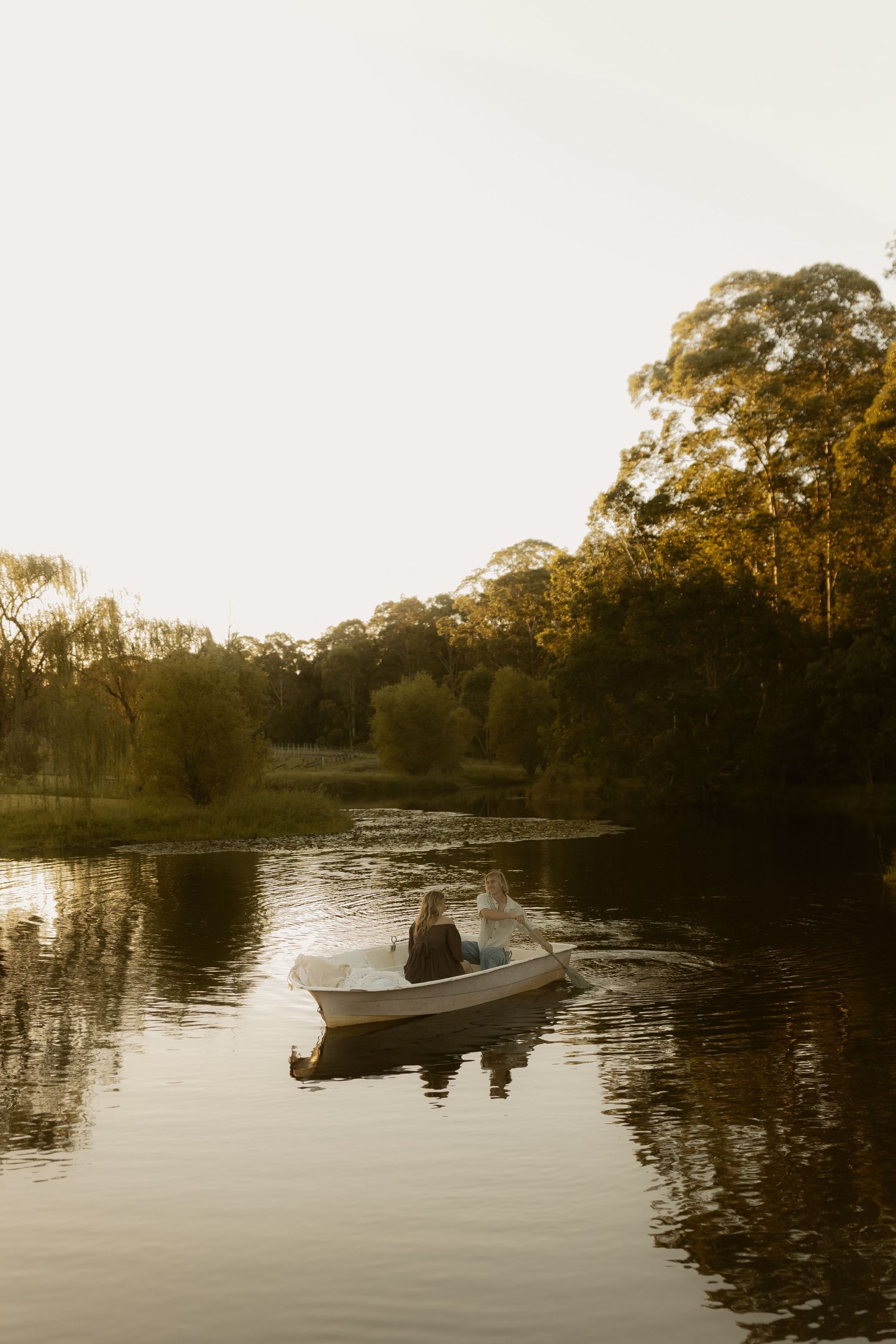 Couple sitting in a white rowboat on a calm reflective lake surrounded by lush green trees at golden hour.