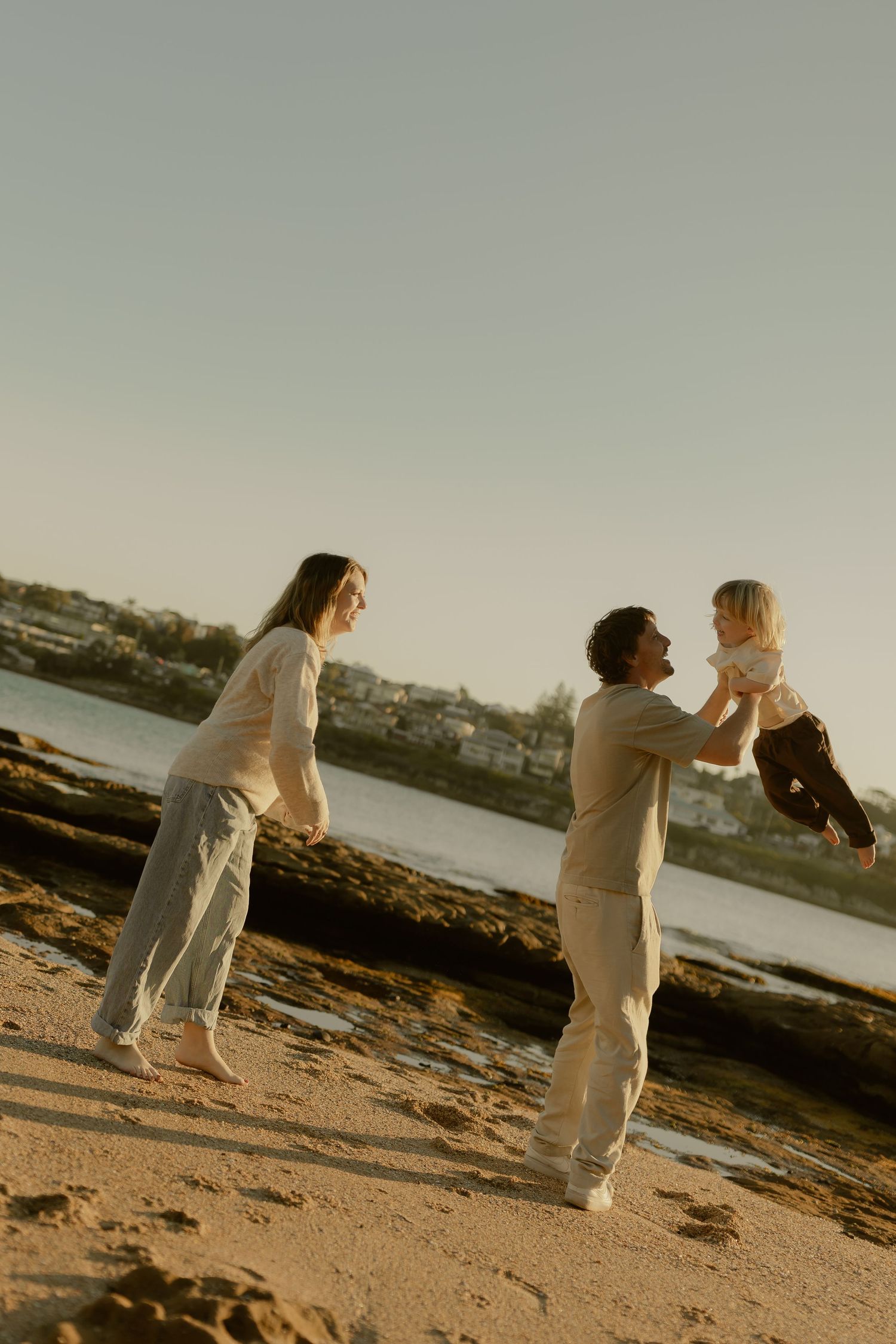 Family playing on a sandy beach at golden hour, father lifting baby while mother watches and smiles nearby.