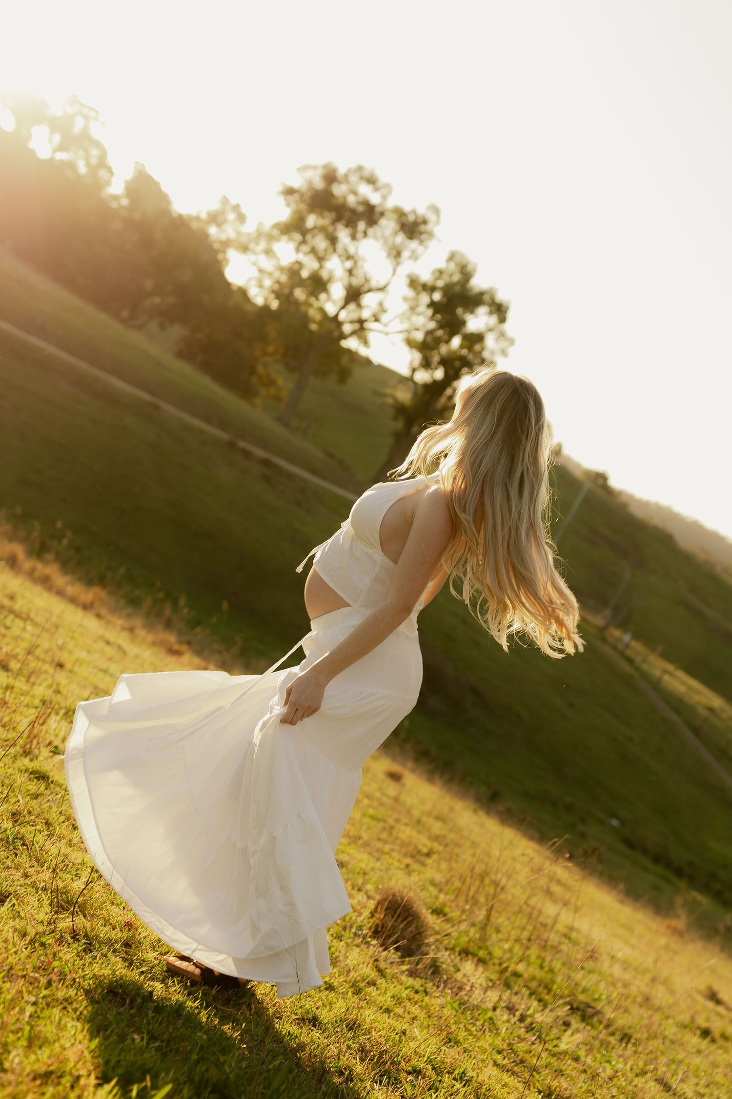 Pregnant woman in flowing white dress twirling in sunlit green hillside meadow.