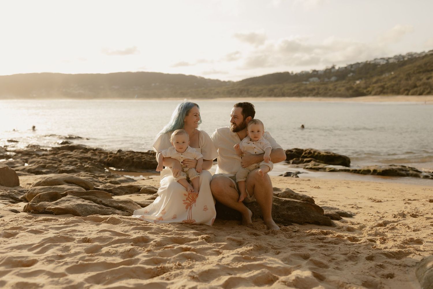A couple shares romantic moments on a rocky beach at sunset with waves crashing in the background.