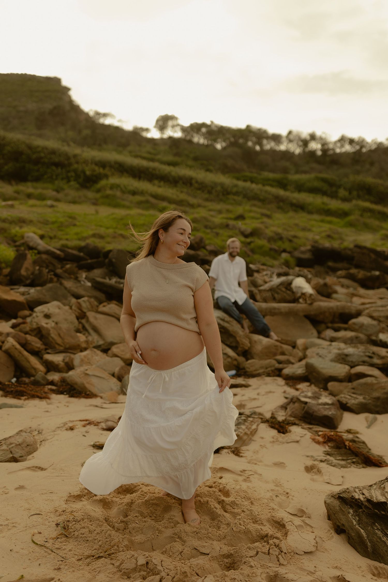 A woman in white dress sits peacefully on coastal rocks during a serene beach photoshoot.