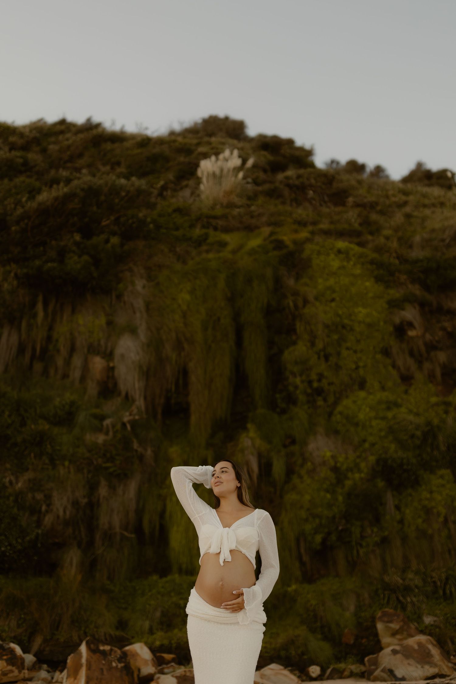 A cliffside maternity session featuring dramatic rocky formations and lush greenery in the background.