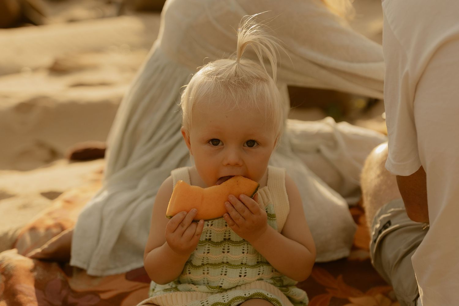 A toddler in a green patterned swimsuit sits on a sandy beach.