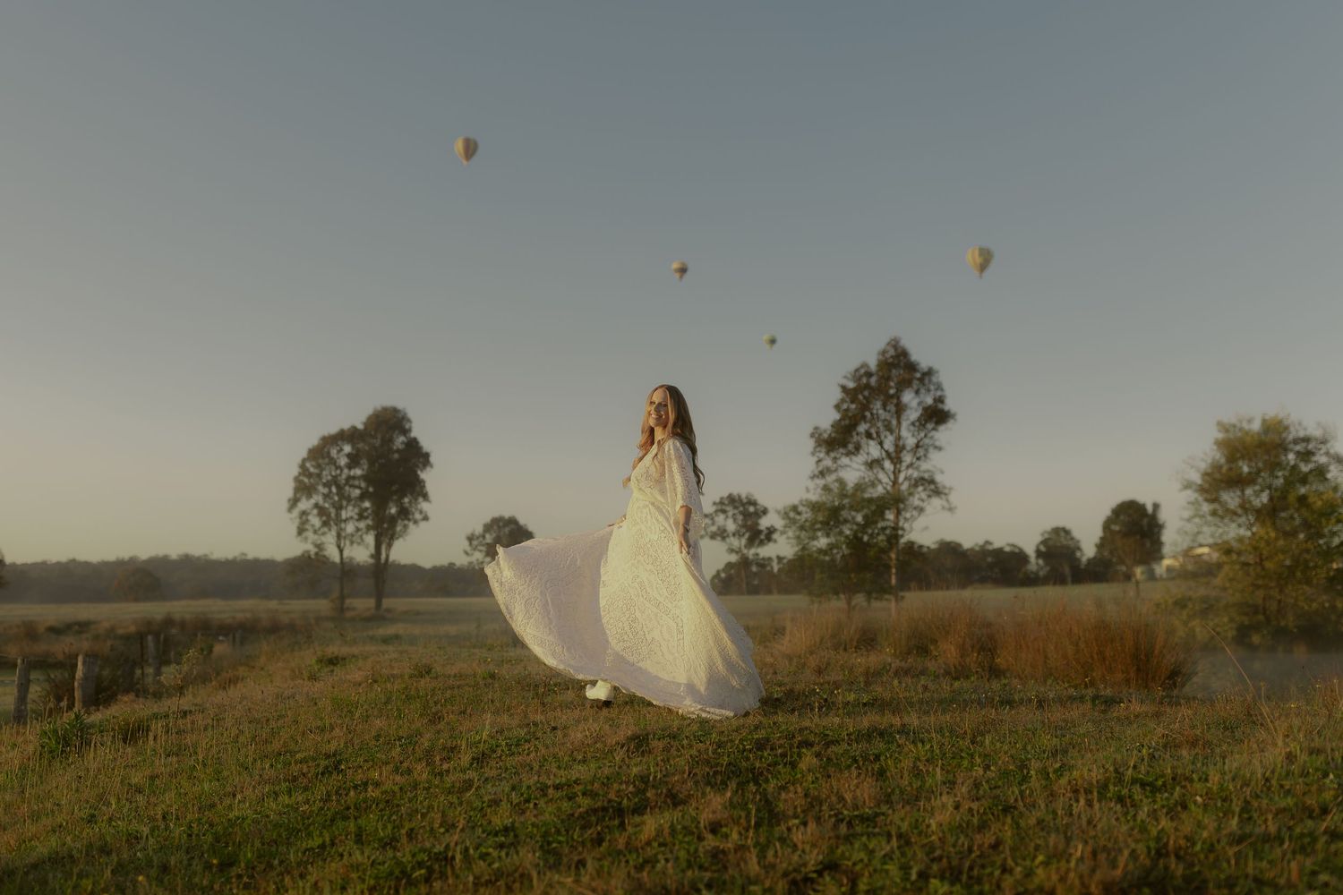 Woman in white flowing dress stands in misty field at dusk with hot air balloons floating in golden sky.