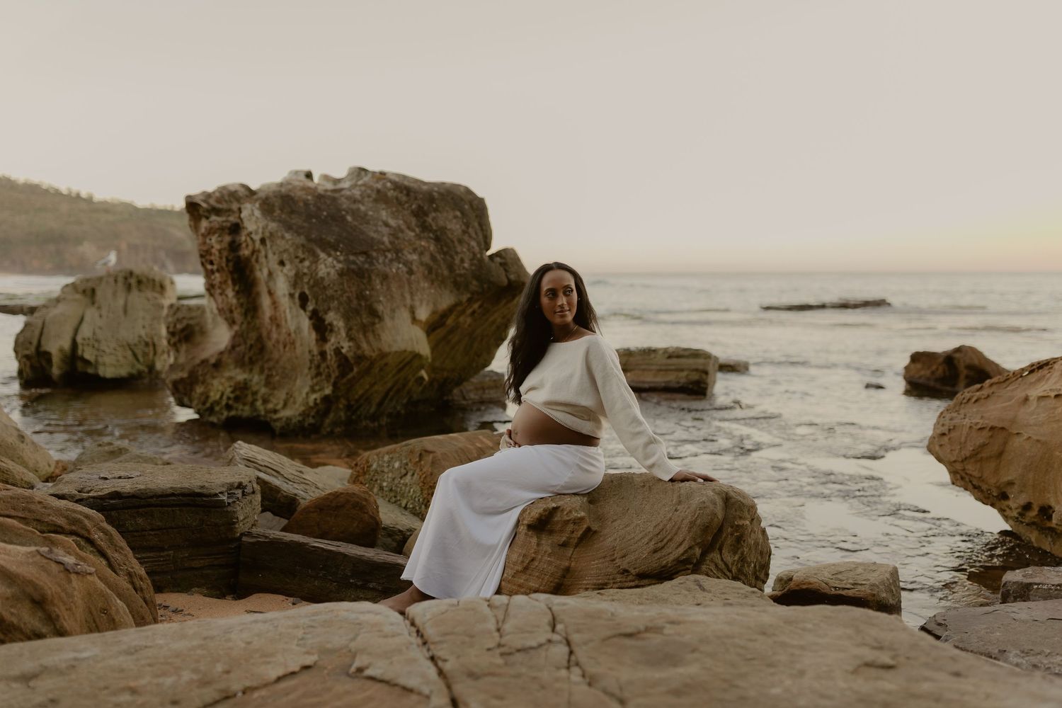 Pregnant woman in white outfit sitting on coastal rocks by the ocean at dusk.