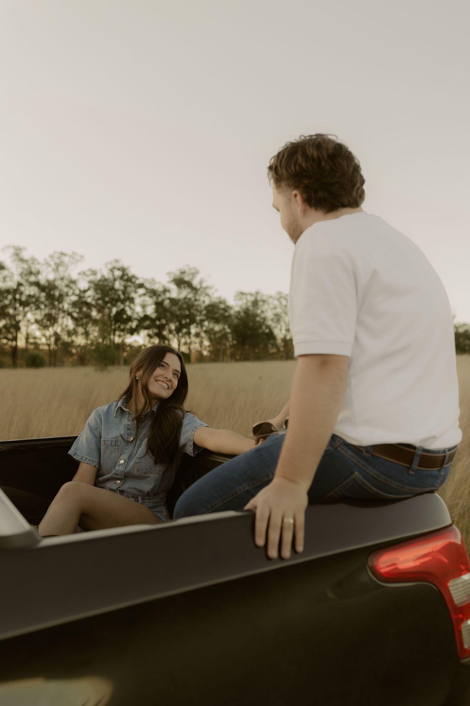 Couple sitting on truck bed in field at sunset, woman smiling at man in white shirt.