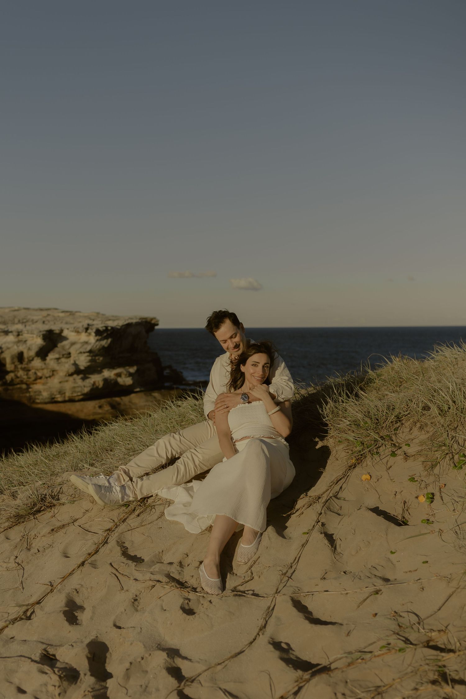 Couple sitting together on sandy coastal dunes at golden hour, with ocean and rocky cliffs in the background.