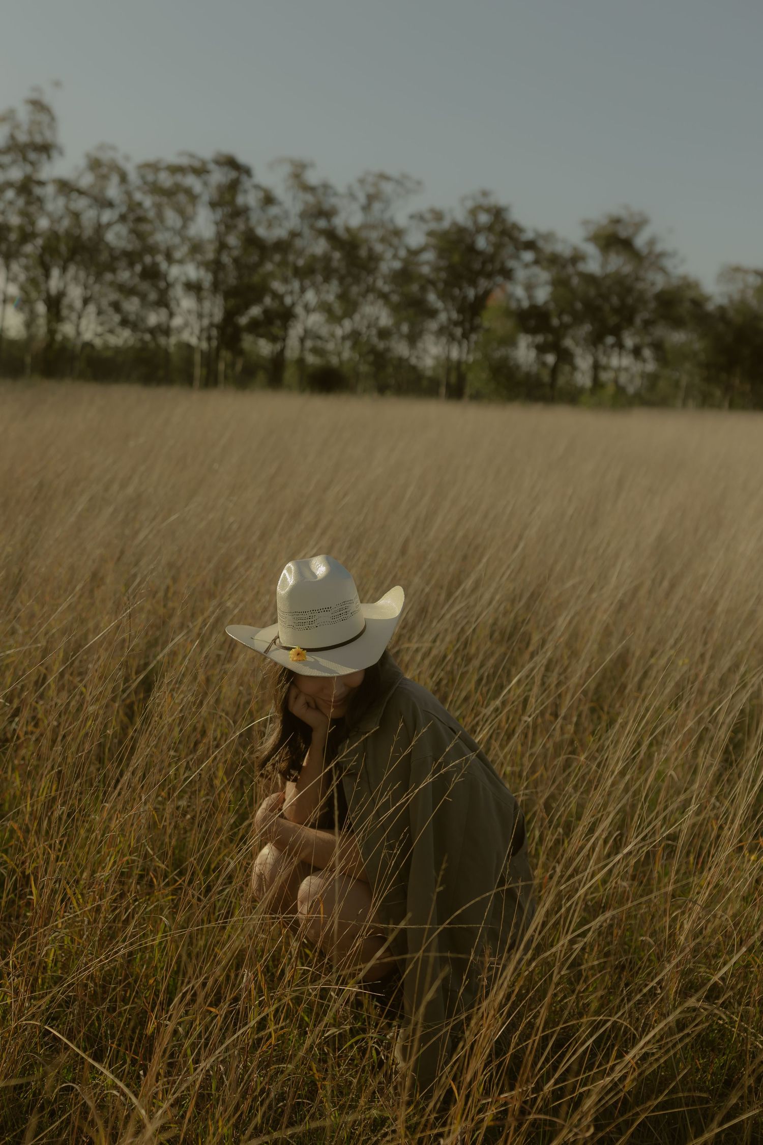 A woman in a white cowboy hat crouches in tall golden grass field under a clear sky.