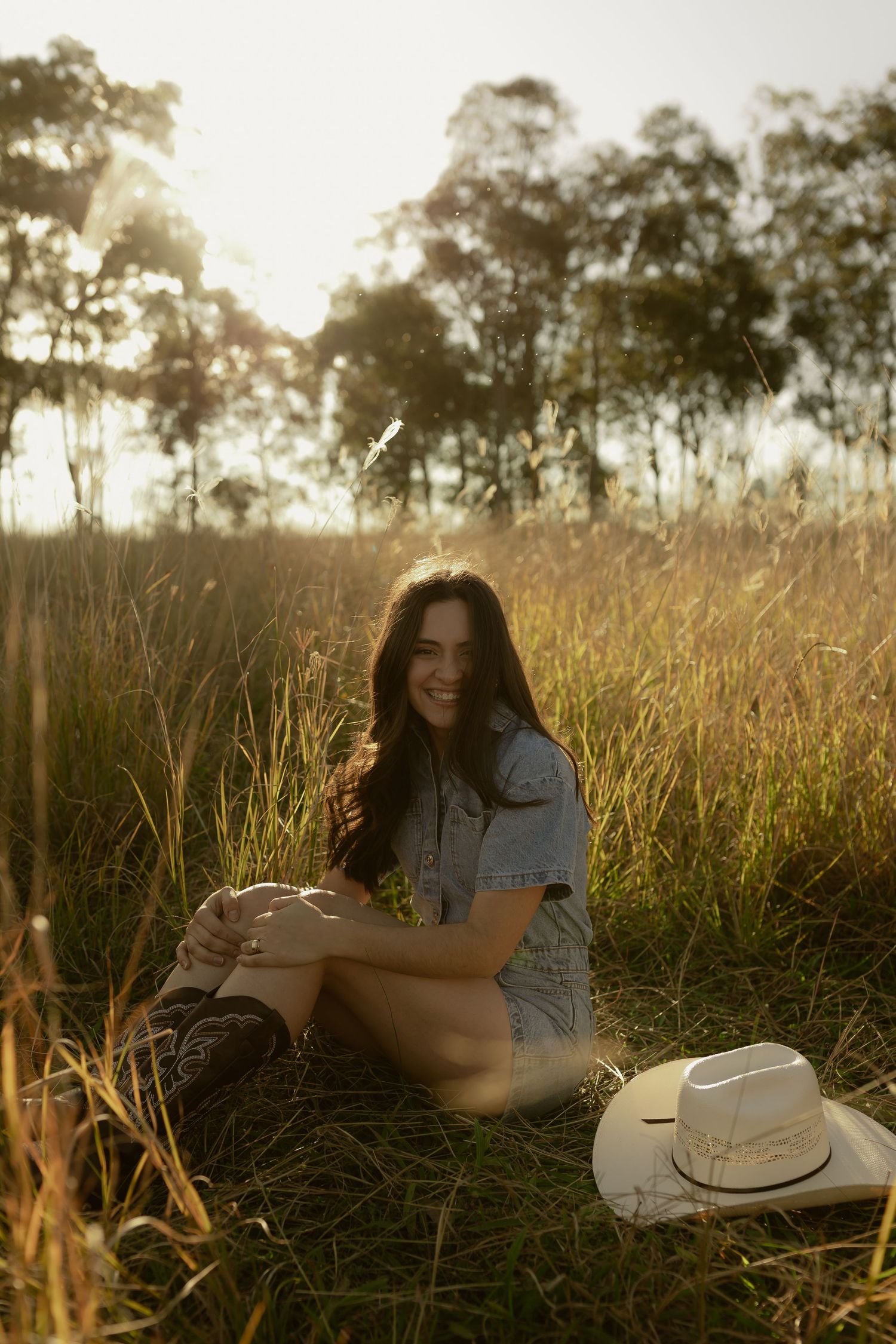 Woman in denim dress sits in golden field at sunset with white cowboy hat.
