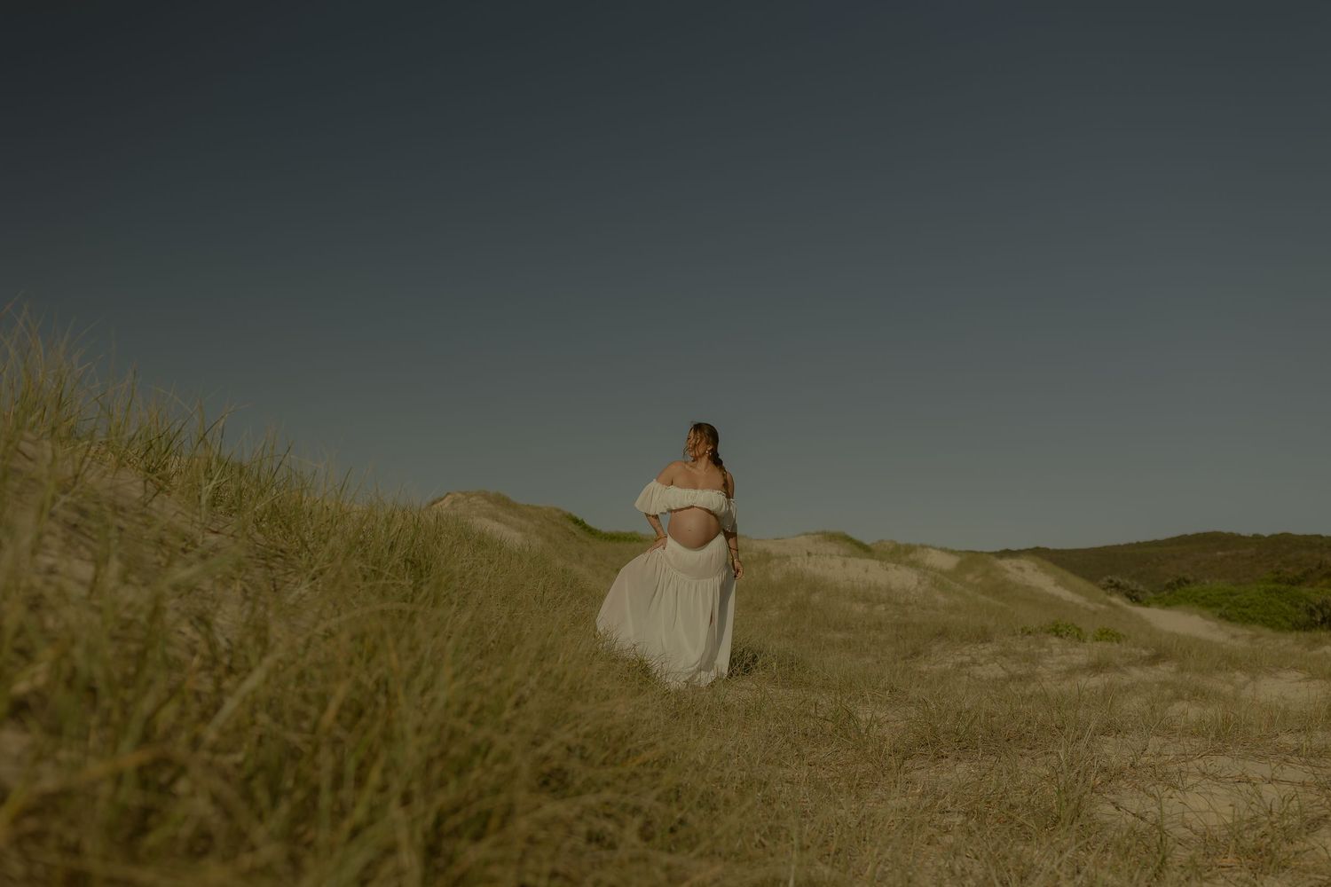 Pregnant woman in white dress standing on sandy dunes under a clear blue sky.