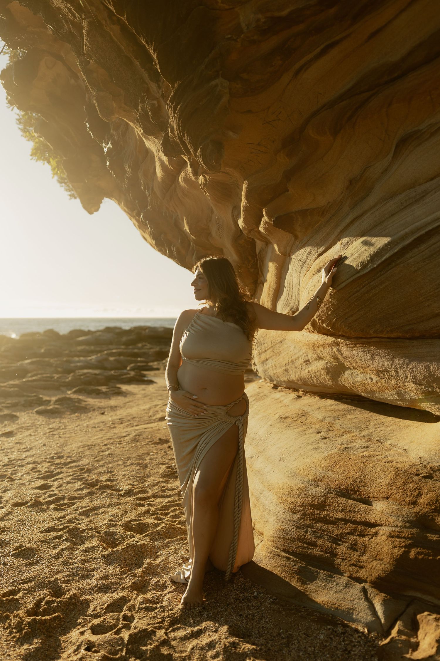 Pregnant woman in flowing white dress touching golden sandstone rock formation on sandy beach at sunset.