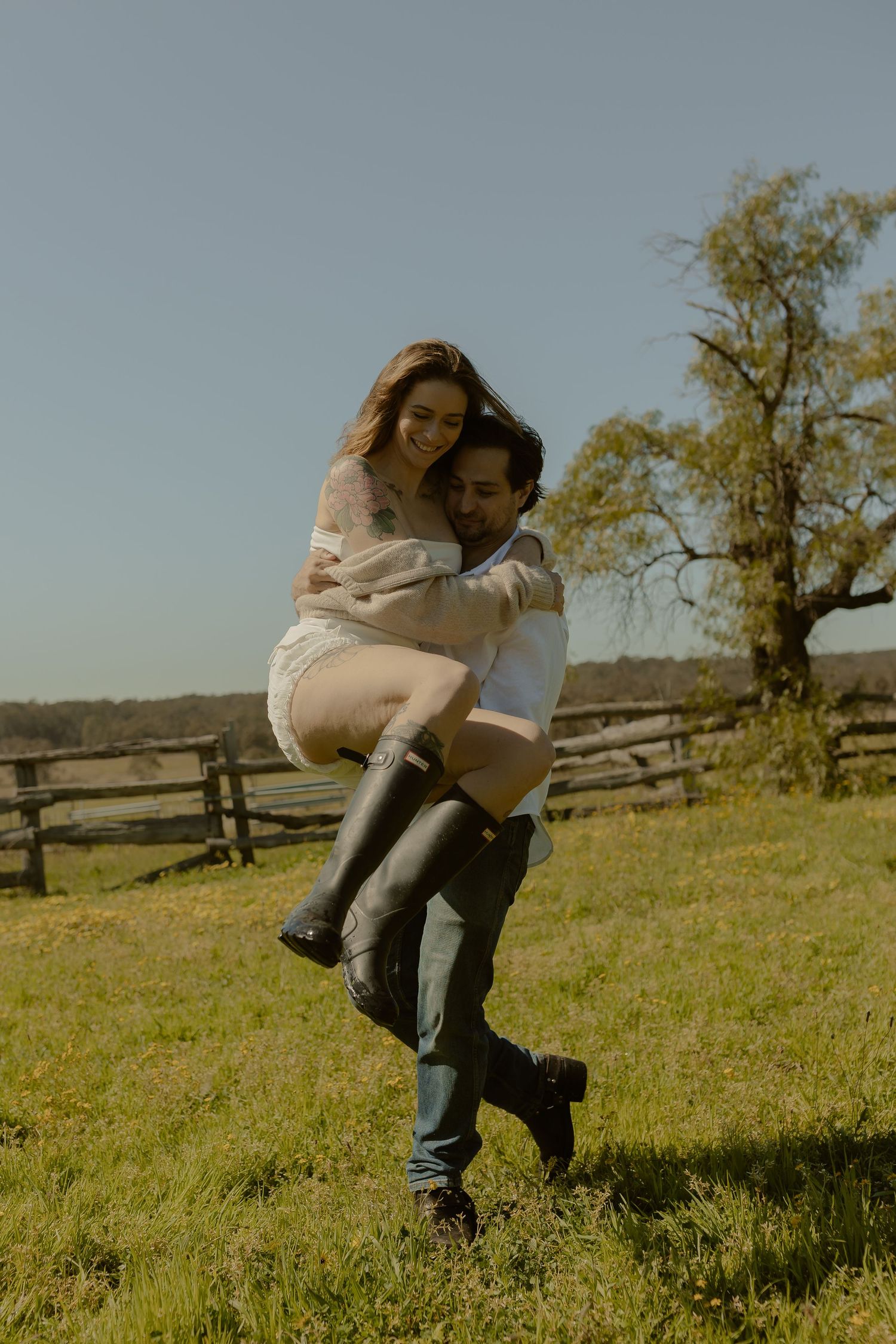 Joyful couple embracing in a sunny field with wooden fence in background.