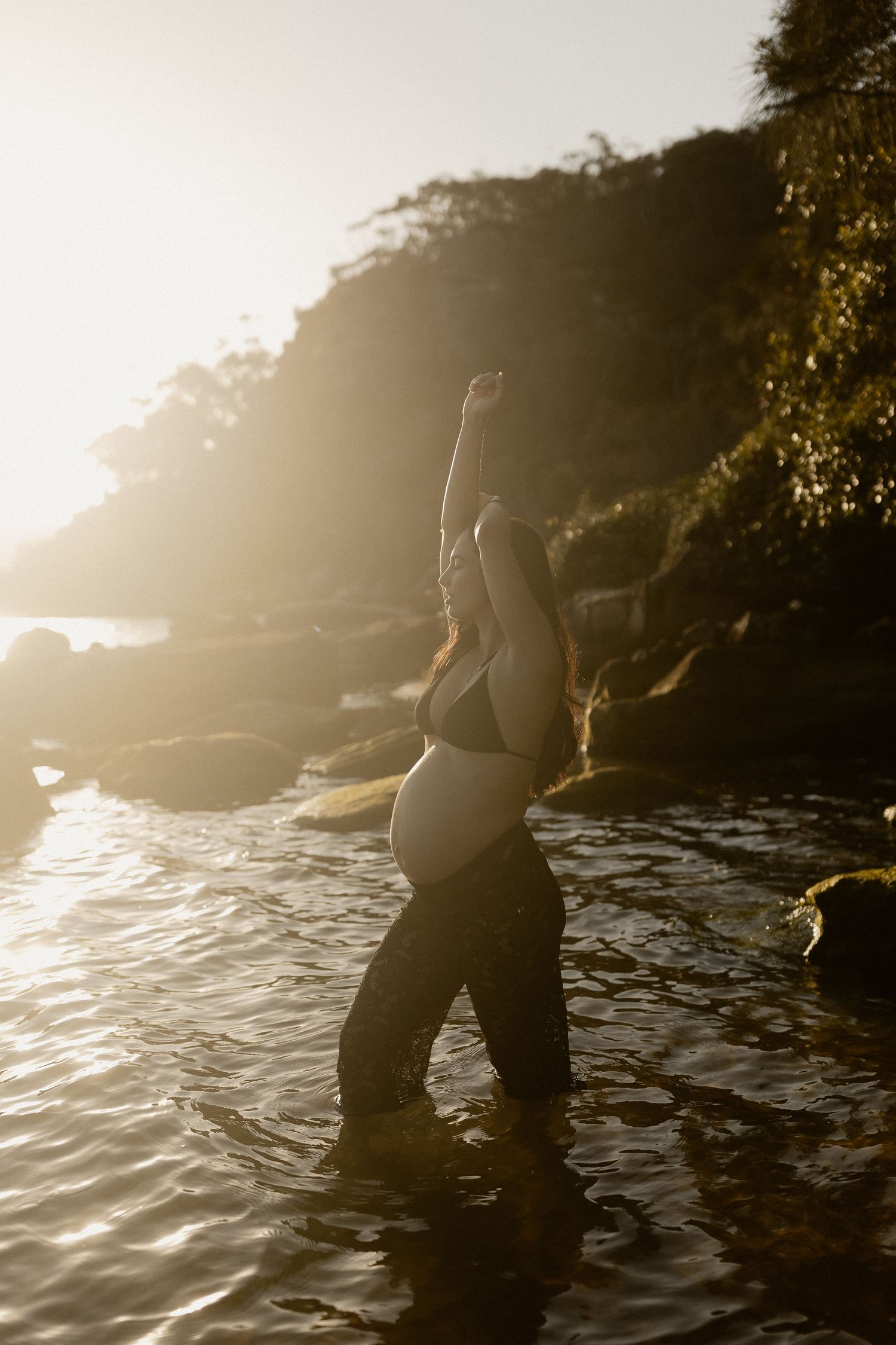 Silhouette of a pregnant figure against sunlit water and rocky coastline at golden hour.