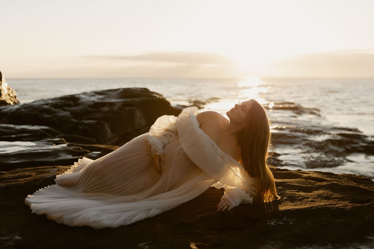 A white swan stretches its elegant wings while perched by the ocean at sunset with light glowing through its feathers.