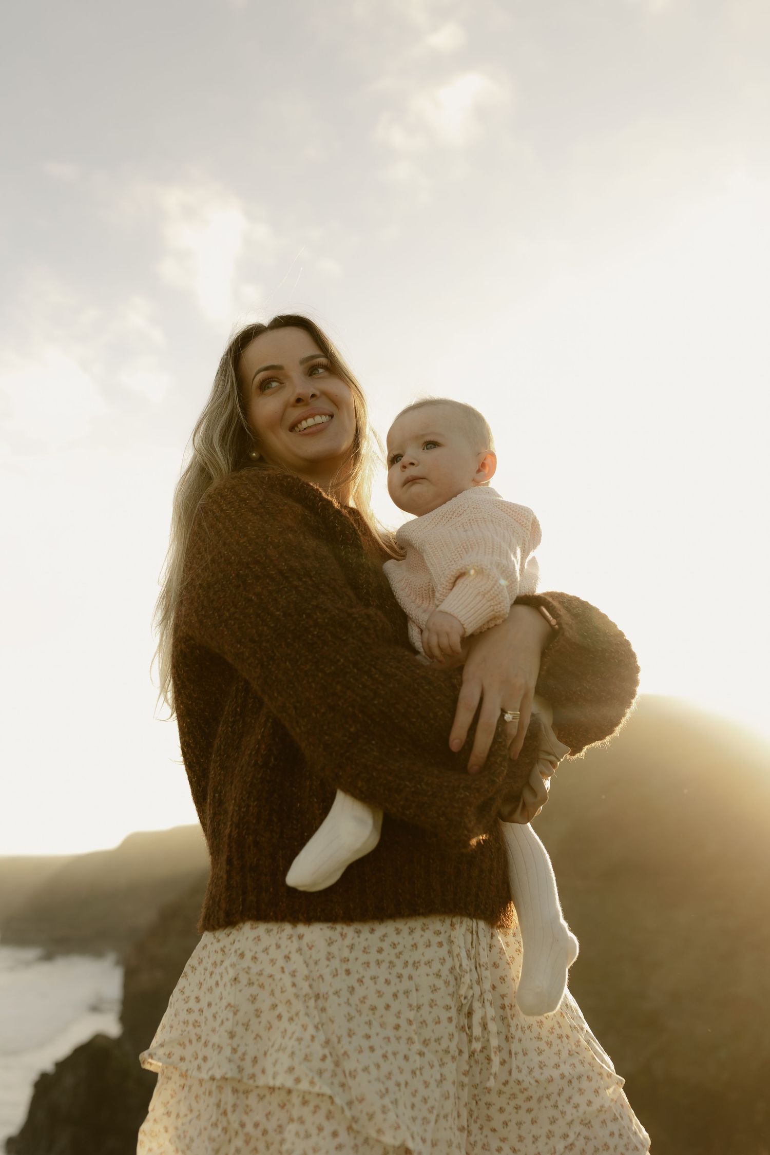 Smiling mother holding baby at sunset on coastal cliffs.